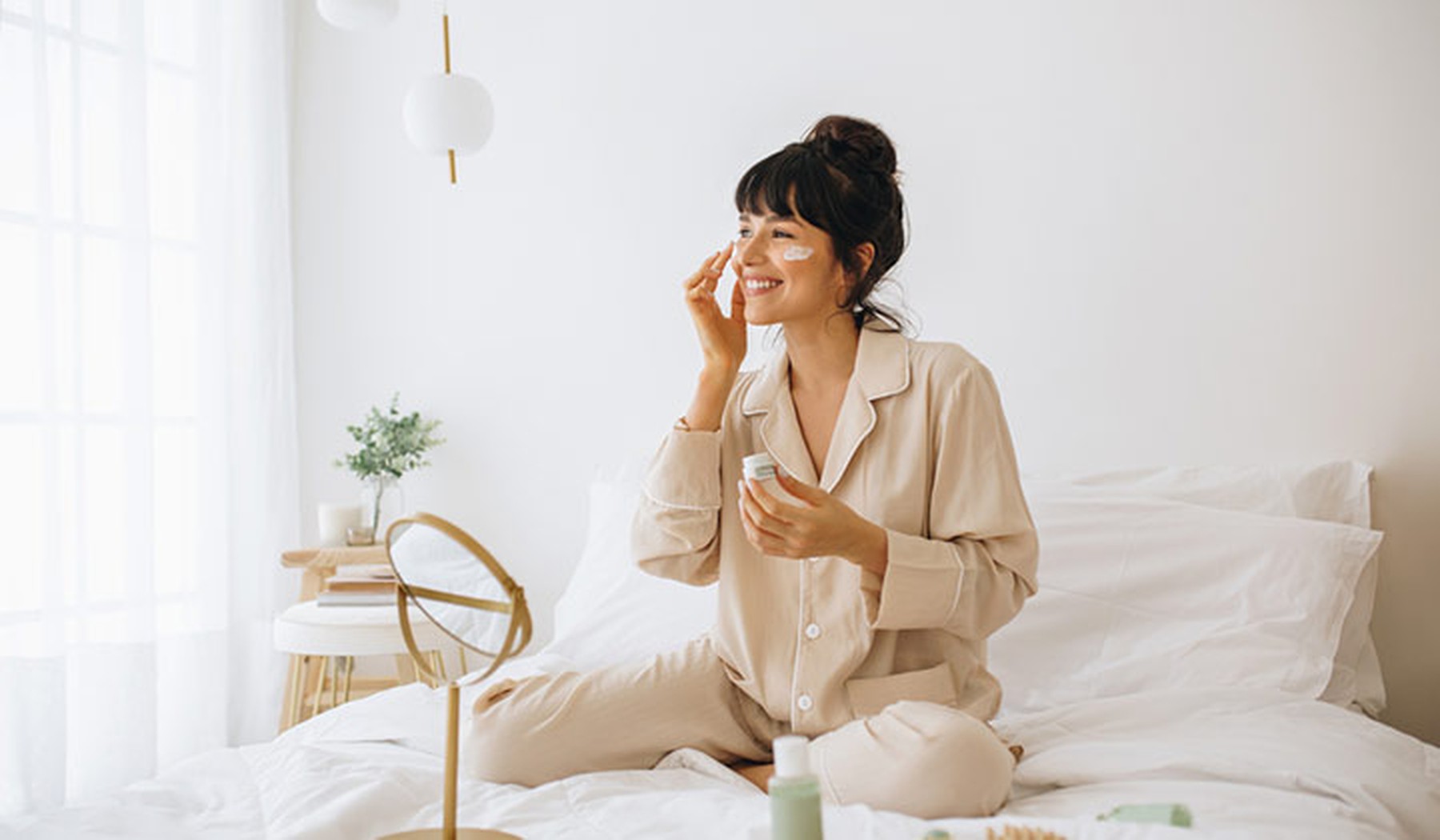 mujer joven sentada en una cama blanca y echándose crema en la cara con un espejo redondo delante
