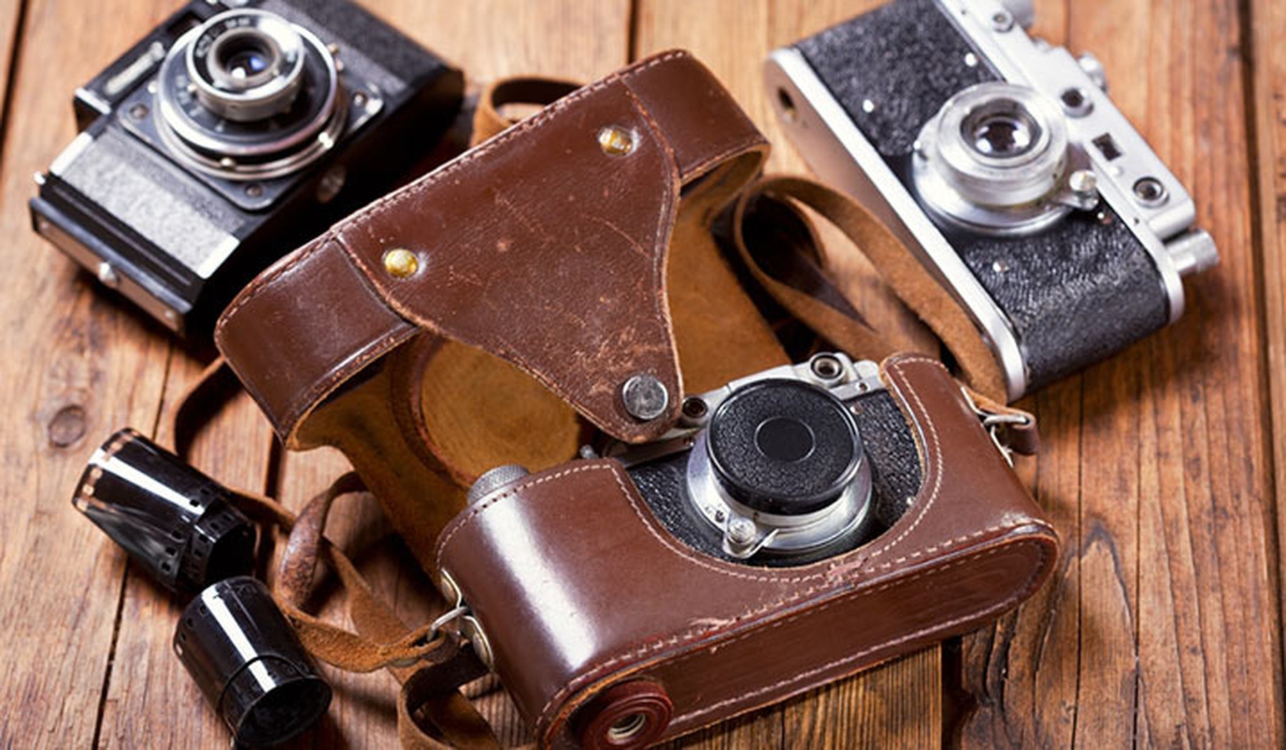 3 cámaras de fotos vintage encima de una mesa de madera. La que está en primer plano tiene una funda marrón de cuero abierta. También hay unos rollos de carrete