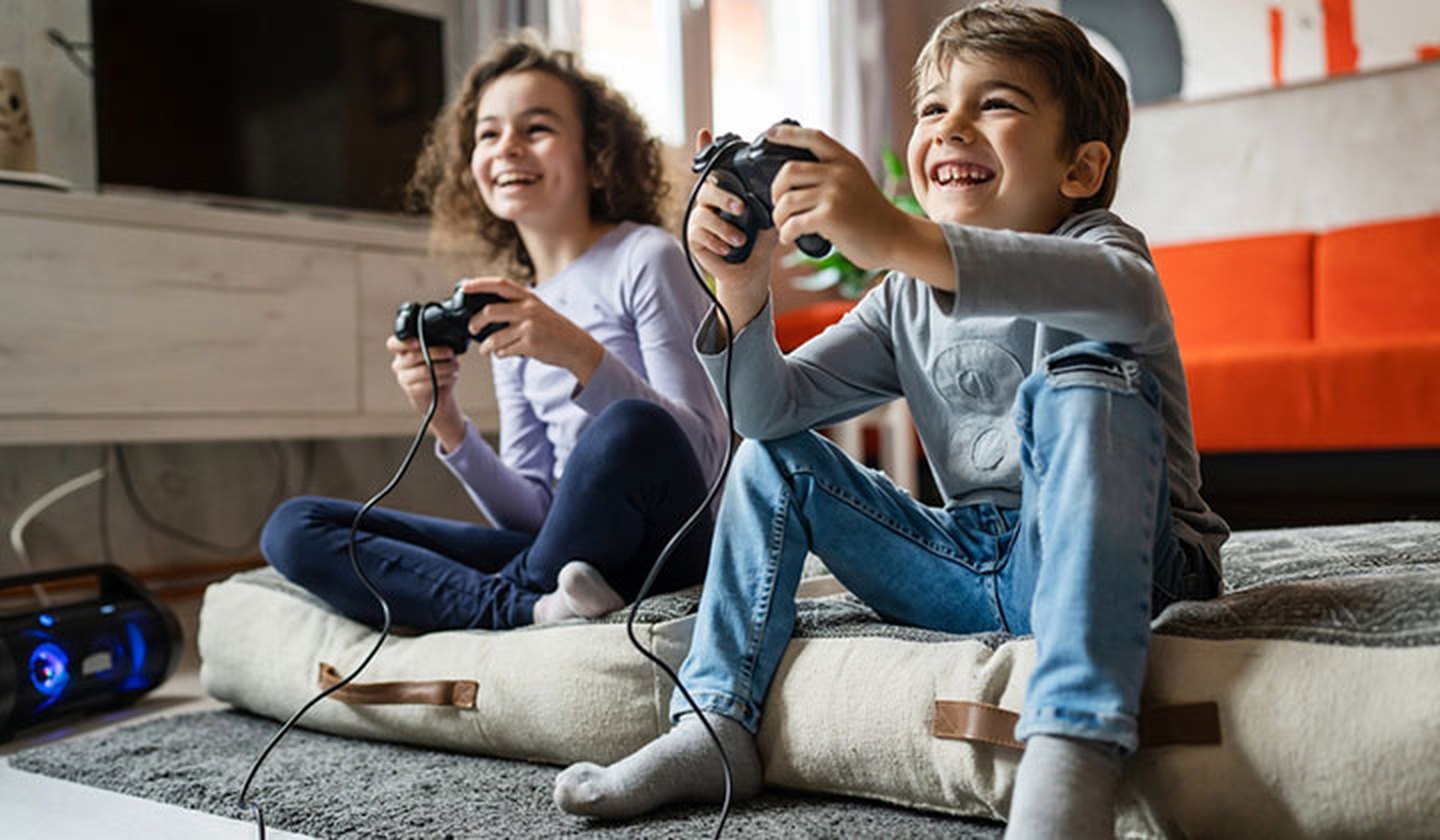 Un niño y una niña sonrientes jugando a videojuegos mientras están sentados en un cojín en el suelo de un salón