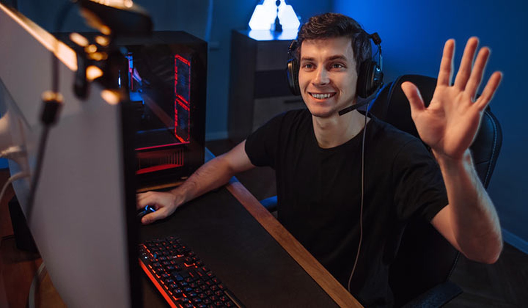 Chico joven sentado frente al ordenador y saludando a una webcam. Lleva unos cascos de gamer y está sentado en una silla de escritorio negra.