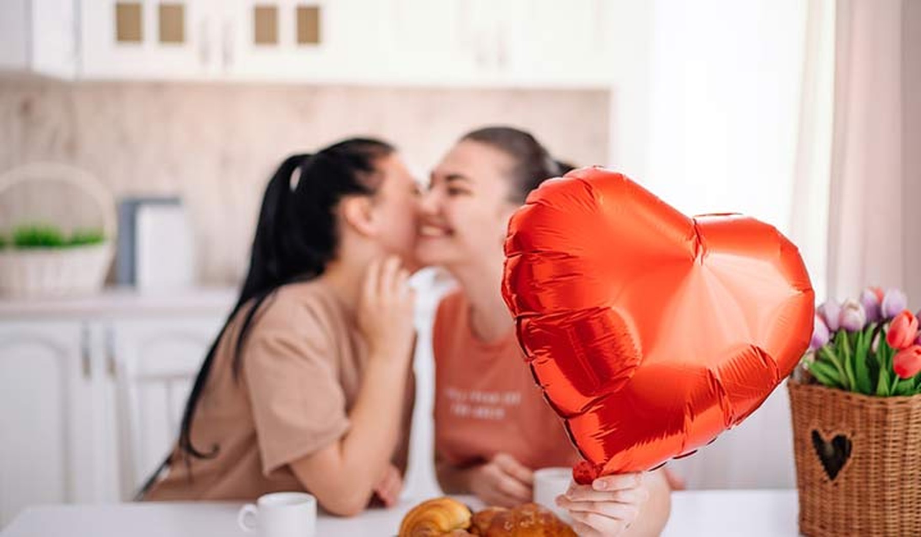 Una pareja joven de dos mujeres dándose un beso en la mejilla mientras desayunan en su cocina. Una de ellas sujeta en primer plano un globo en forma de corazón.