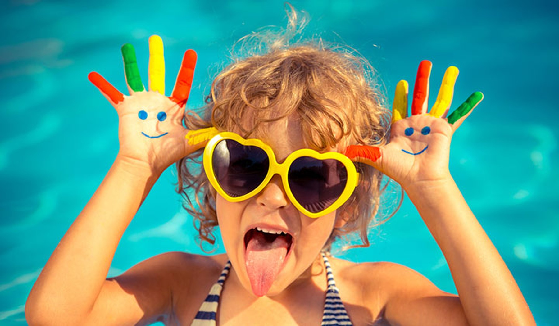 Una niña con el pelo corto y rizado en una piscina con unas gafas de sol amarillas en forma de corazón y las manos pintadas de colores. Tiene las manos a la altura de la cabeza y saca la lengua.