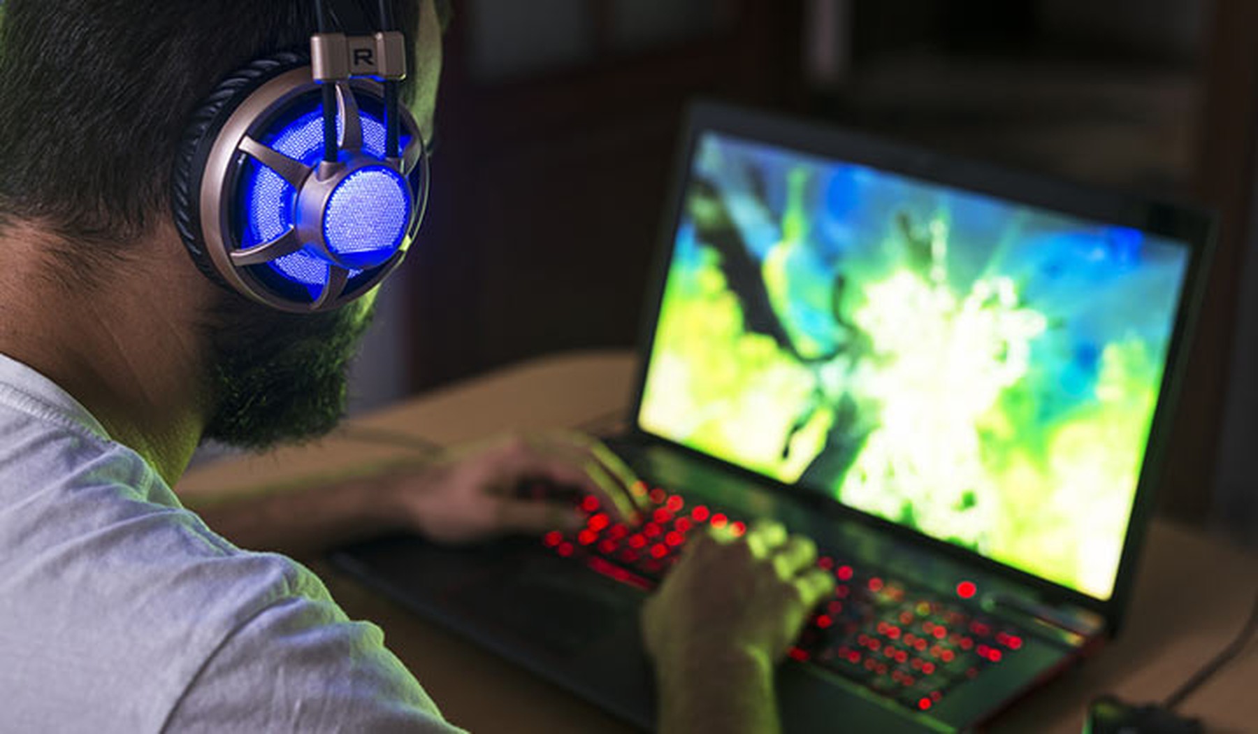 portátil de gaming con las teclas iluminadas en rojo y un vídeojuego en pantalla. Usándolo hay un hombre joven con barba y unos cascos iluminados en azul