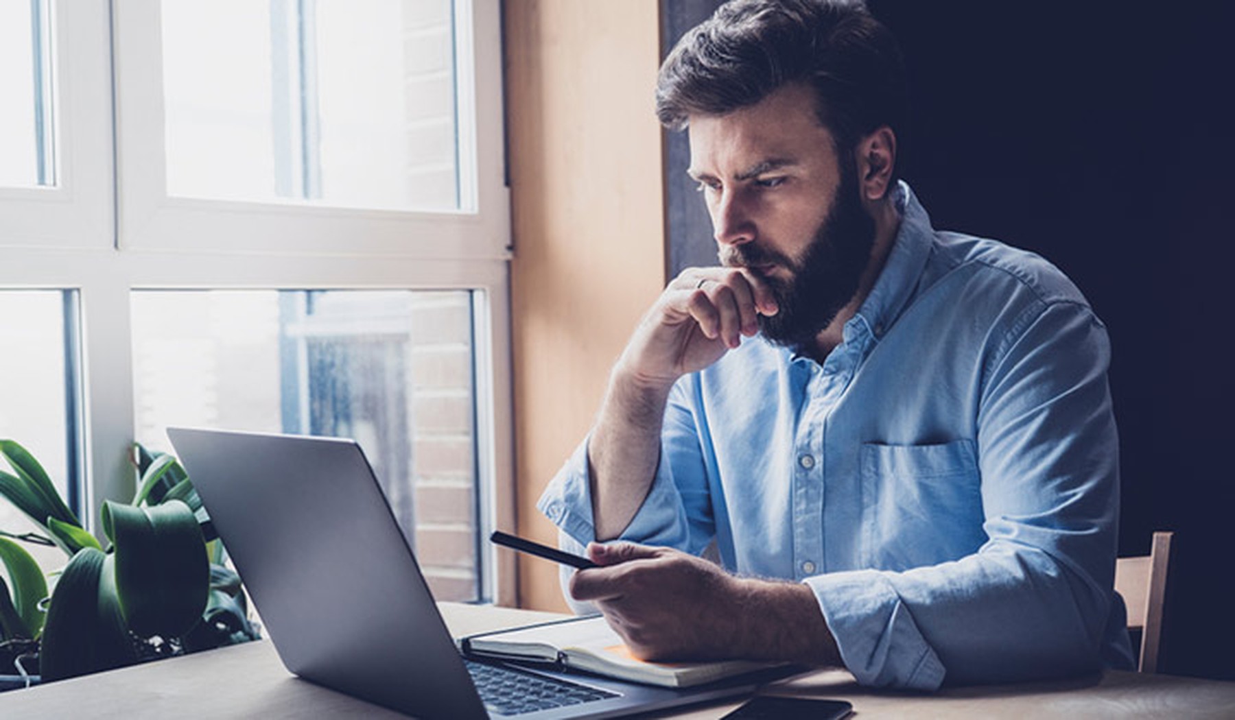 Hombre con camisa azul mirando la pantalla de un portátil y con un bolígrafo en la mano