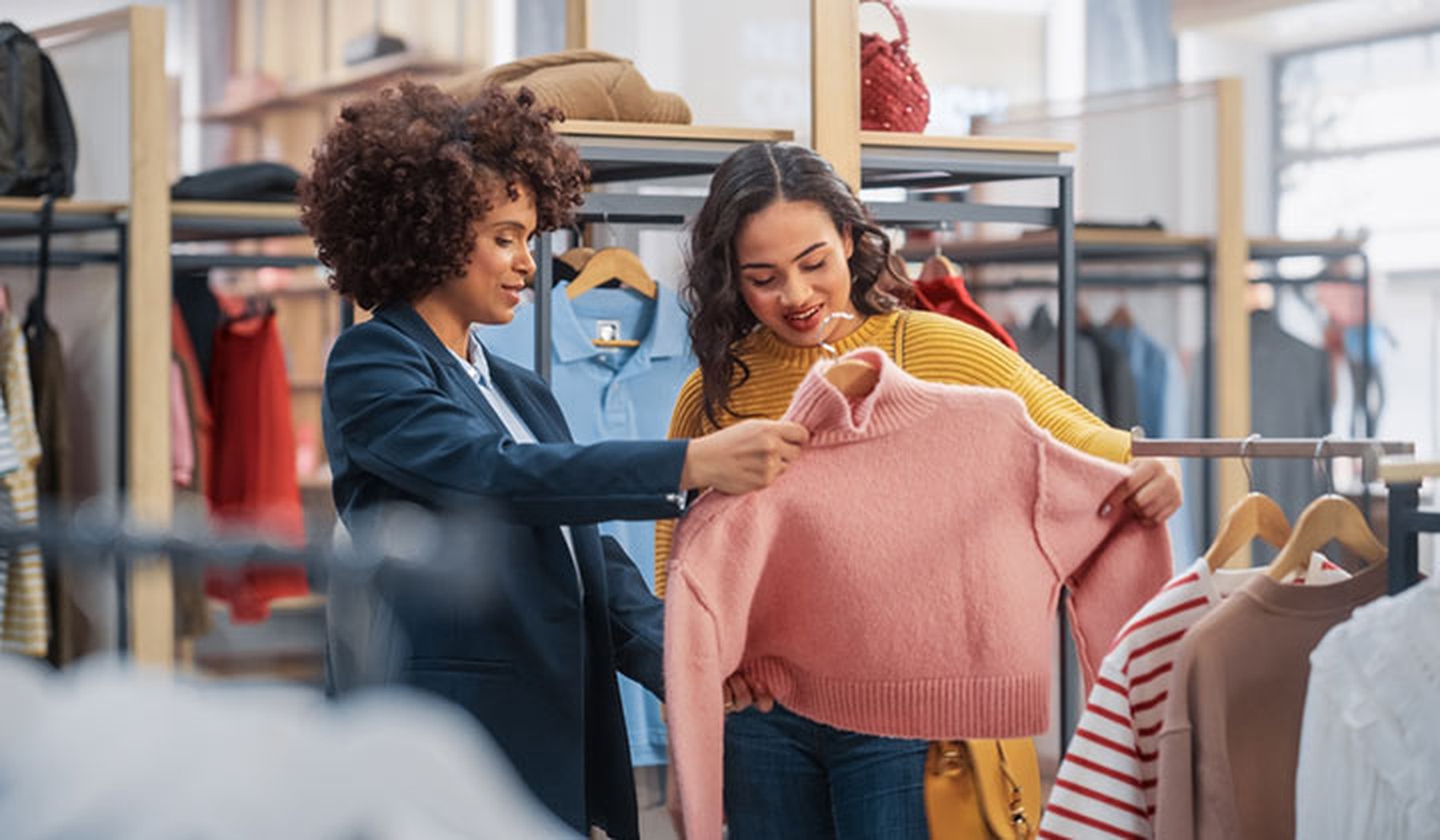 Mujer adulta con el pelo rizado y traje de chaqueta enseñándole un jersey rosa a una mujer joven en una tienda de ropa