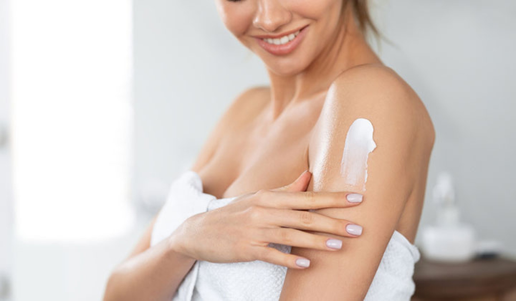 Mujer joven echándose crema aftersun en el brazo después de salir de la ducha envuelta en una toalla blanca