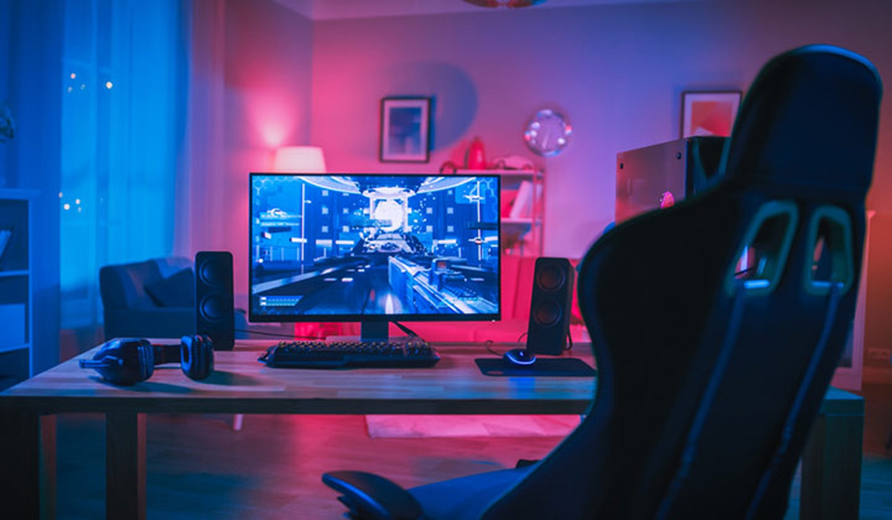 Set de gamer con luces azules y rosas. Silla de gamer en primer plano, monitor con un videojuego puesto encima de una mesa de madera en la que también hay unos cascos y unos altavoces