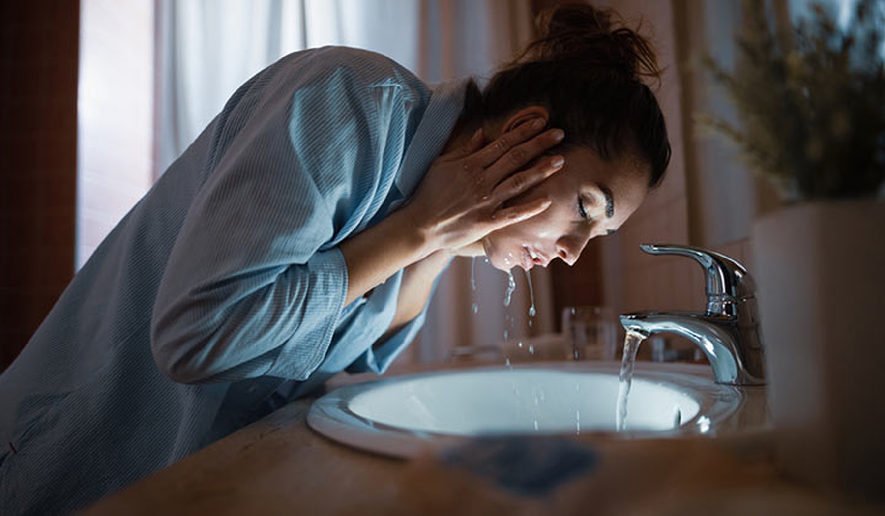 Mujer lavándose la cara en el lavabo de su casa. Lleva un pijama azul y el pelo recogido en un moño.