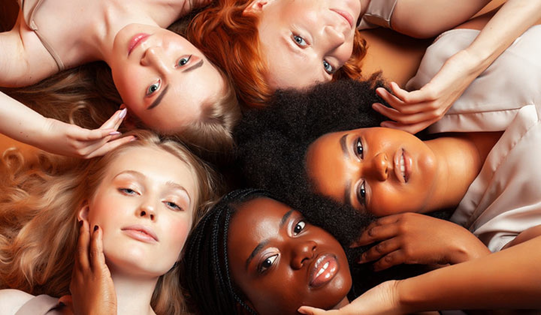5 mujeres tumbadas en círculo con las cabezas tocándose. Cada una tiene un tono de piel diferente. 