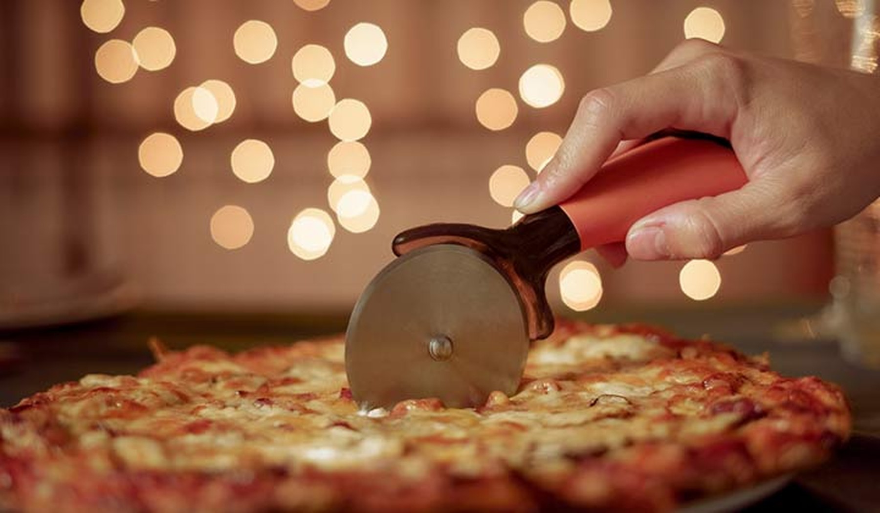 Persona cortando una pizza con un corta pizzas y unas luces de fondo