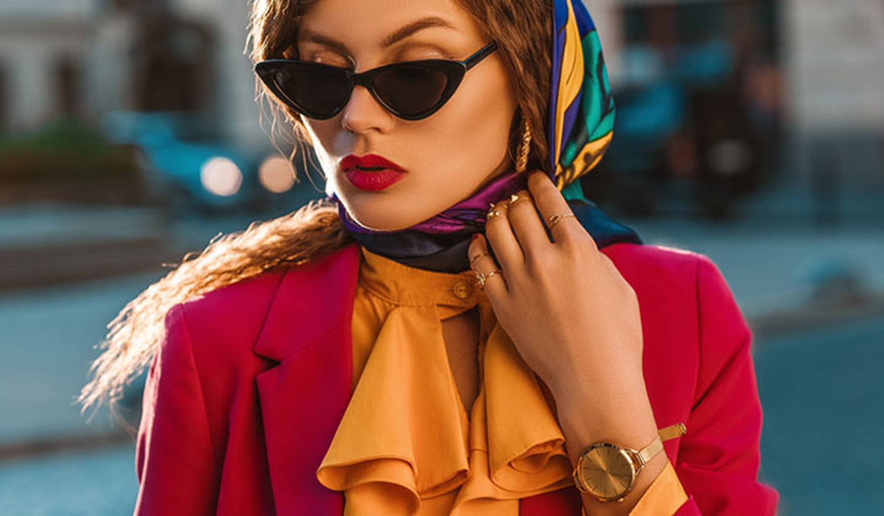 Mujer joven con un pañuelo retro en la cabeza, unas gafas de sol con forma triangular, labios pintados de rojo a juego de su blazer, camisa con cuello con volantes naranjas y accesorios dorados (reloj y anillos)