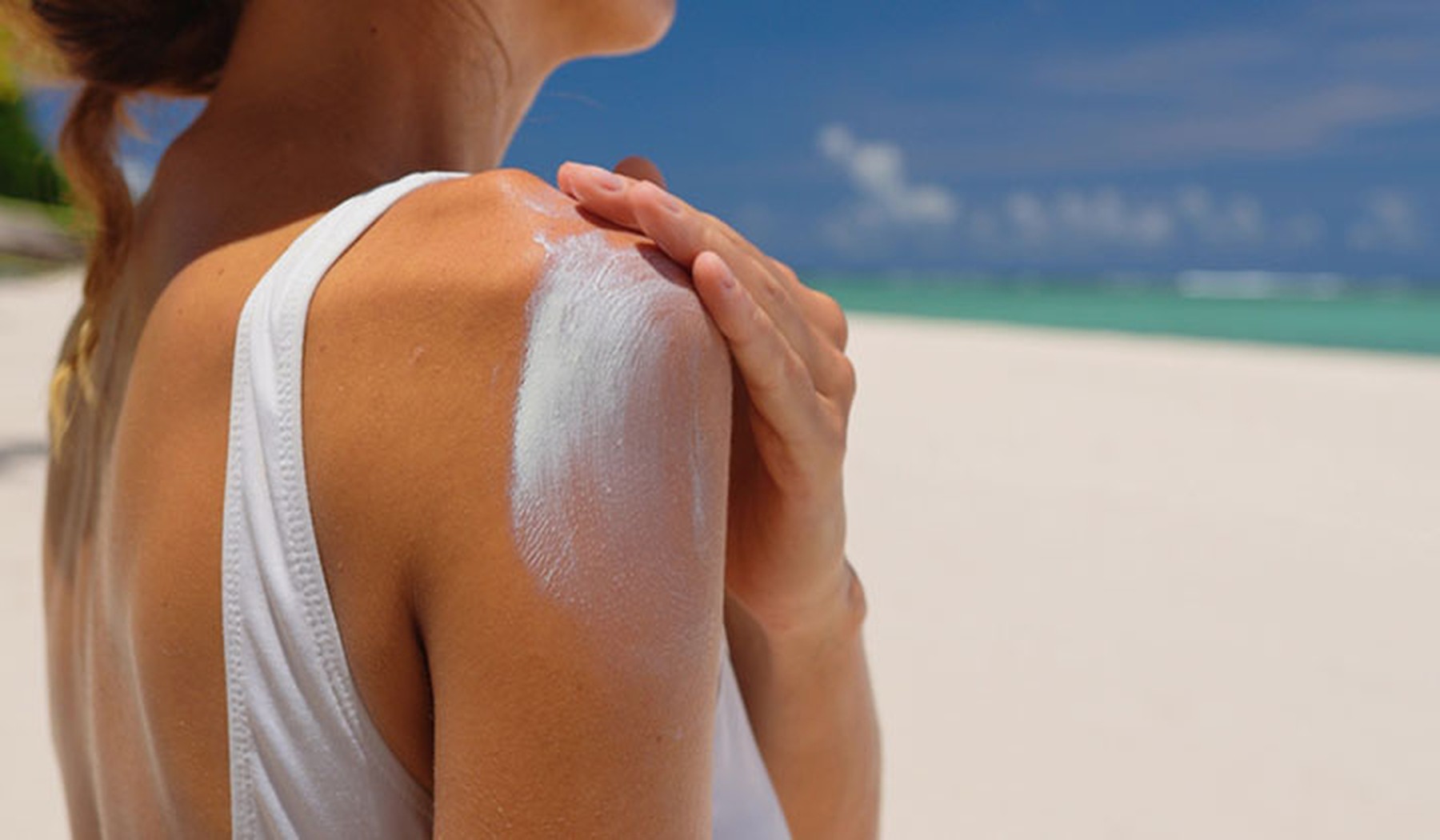 Mujer echándose crema solar en el hombro. Lleva un bañador blanco y de fondo se ve una playa paradisiaca.