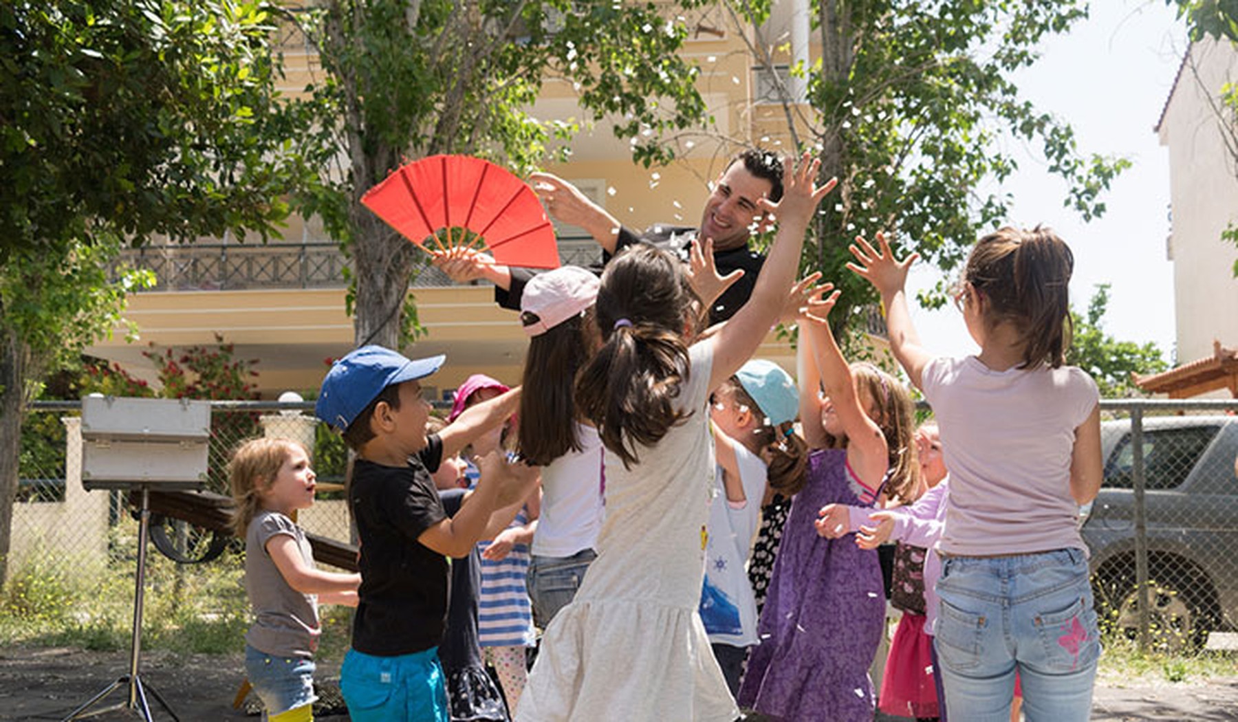 Hombre haciendo un espectáculo en una fiesta de niños. El hombre tiene un abanico rojo en la mano y los niños saltan a su alrededor.