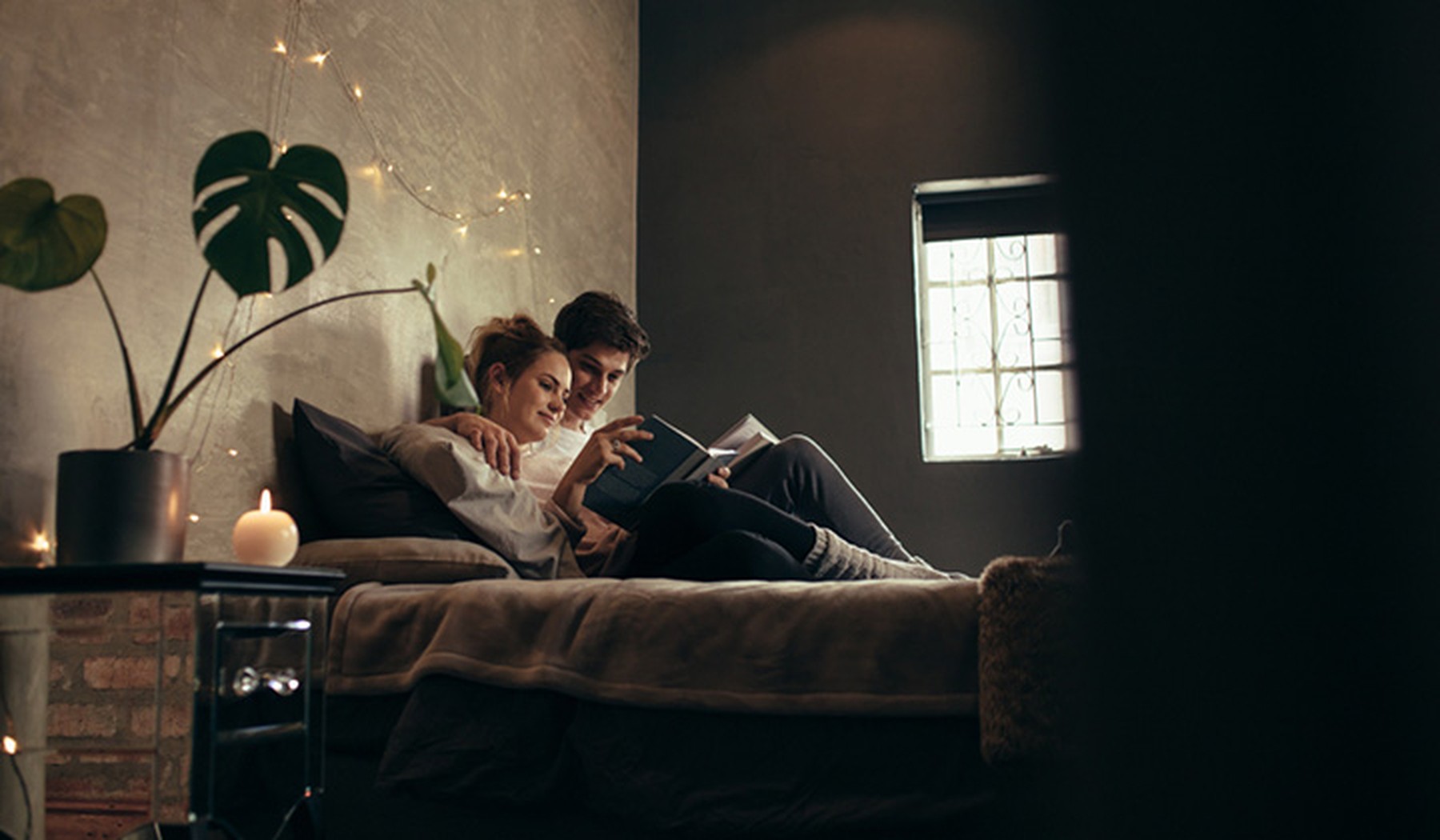 Pareja joven de chico y chica tumbados en una cama leyendo juntos un libro. La habitación está iluminada por una vela y una guirnalda de lucecitas.