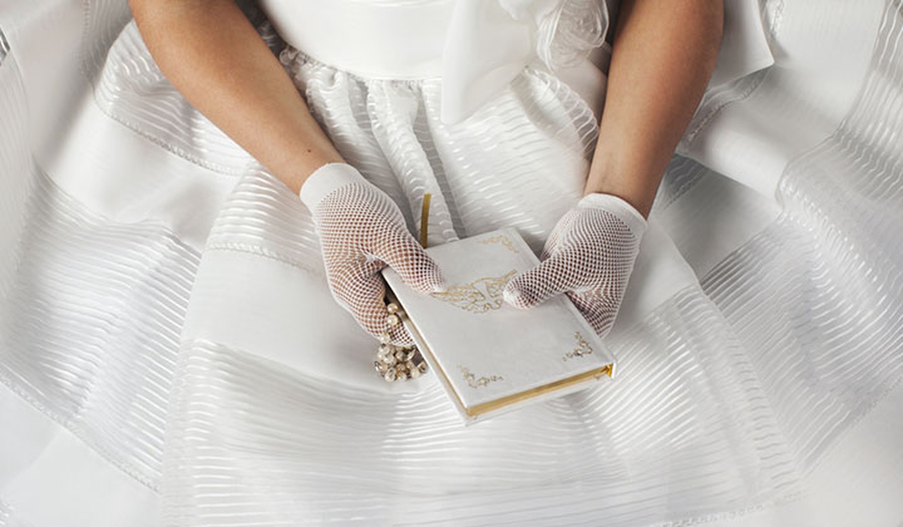 Manos de una niña vestida de comunión sujetando un cuadernito blanco y un rosario. Lleva unos guantes de rejilla blancos.