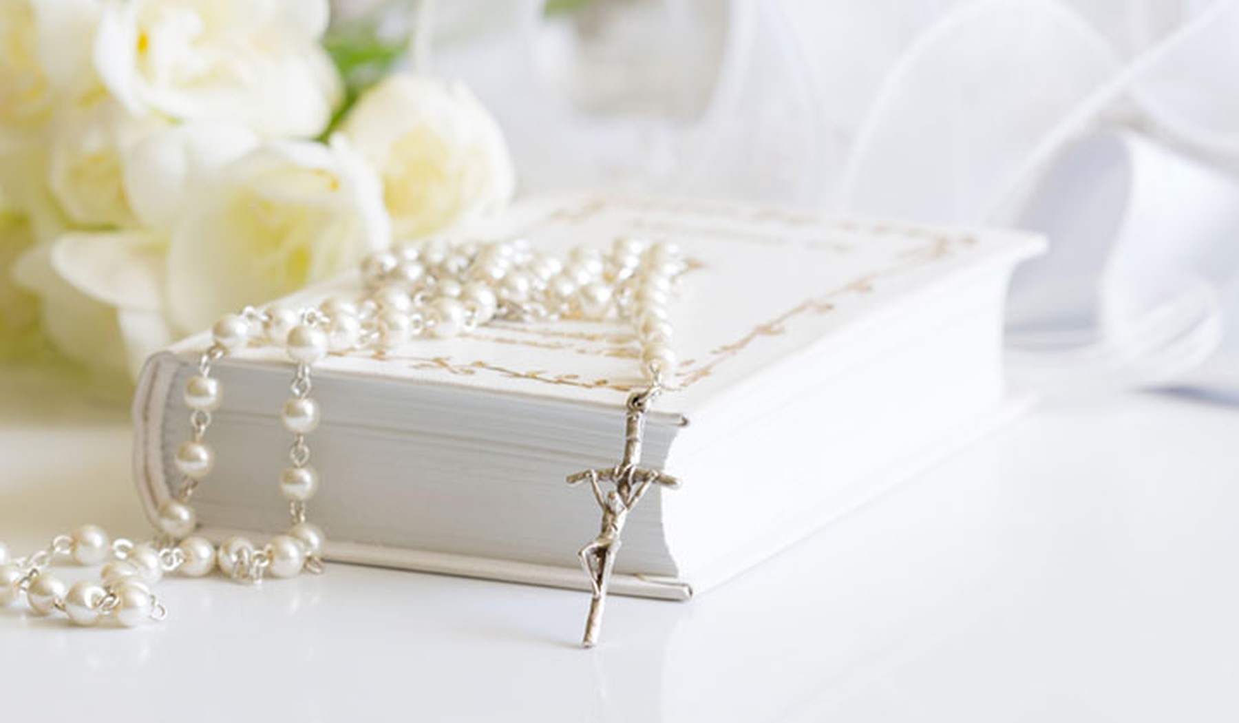 Biblia blanca con un rosario de perlas encima y unas flores blancas de fondo.