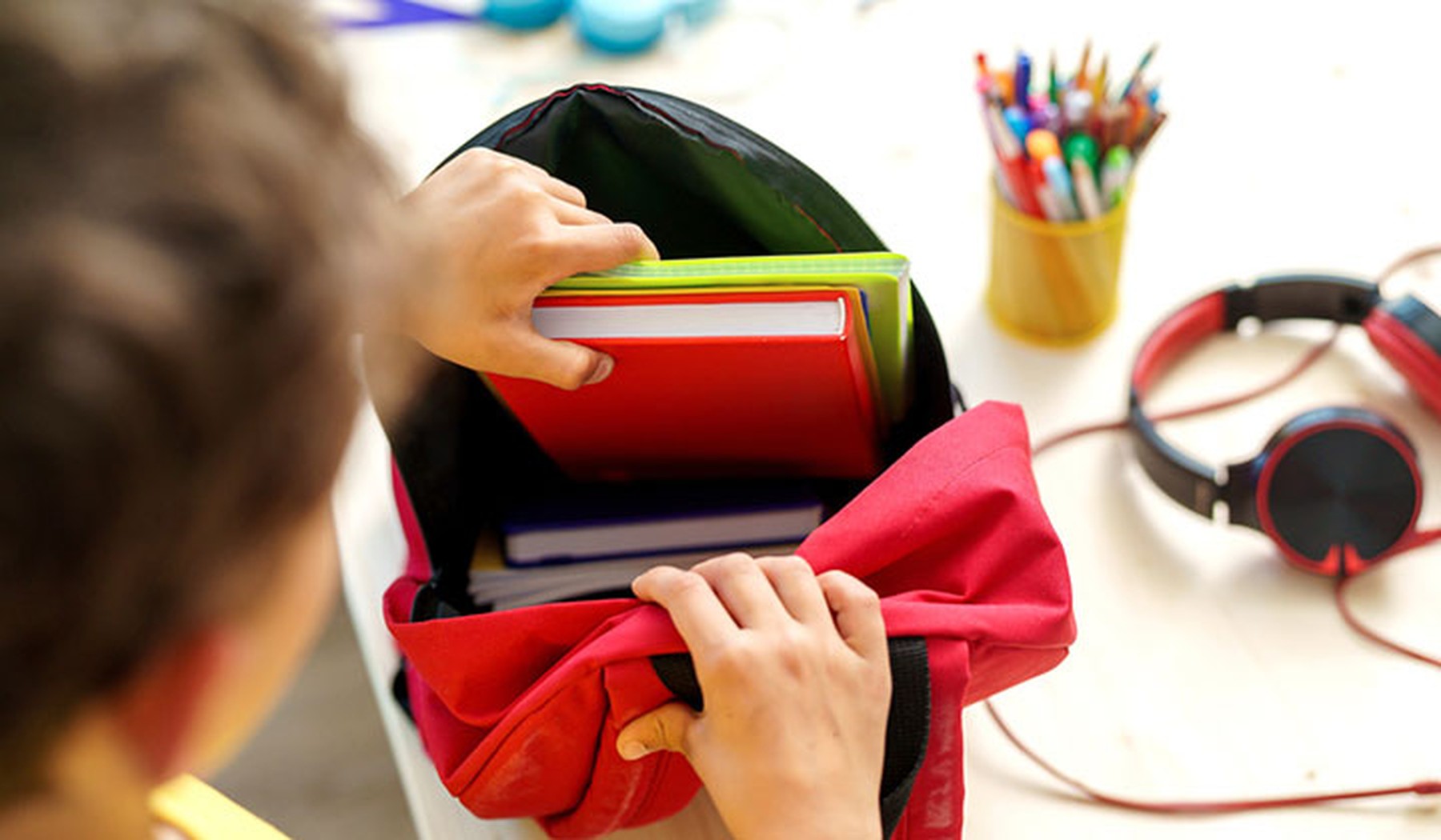 Niño metiendo unos libros rojos y verdes en su mochila escolar roja