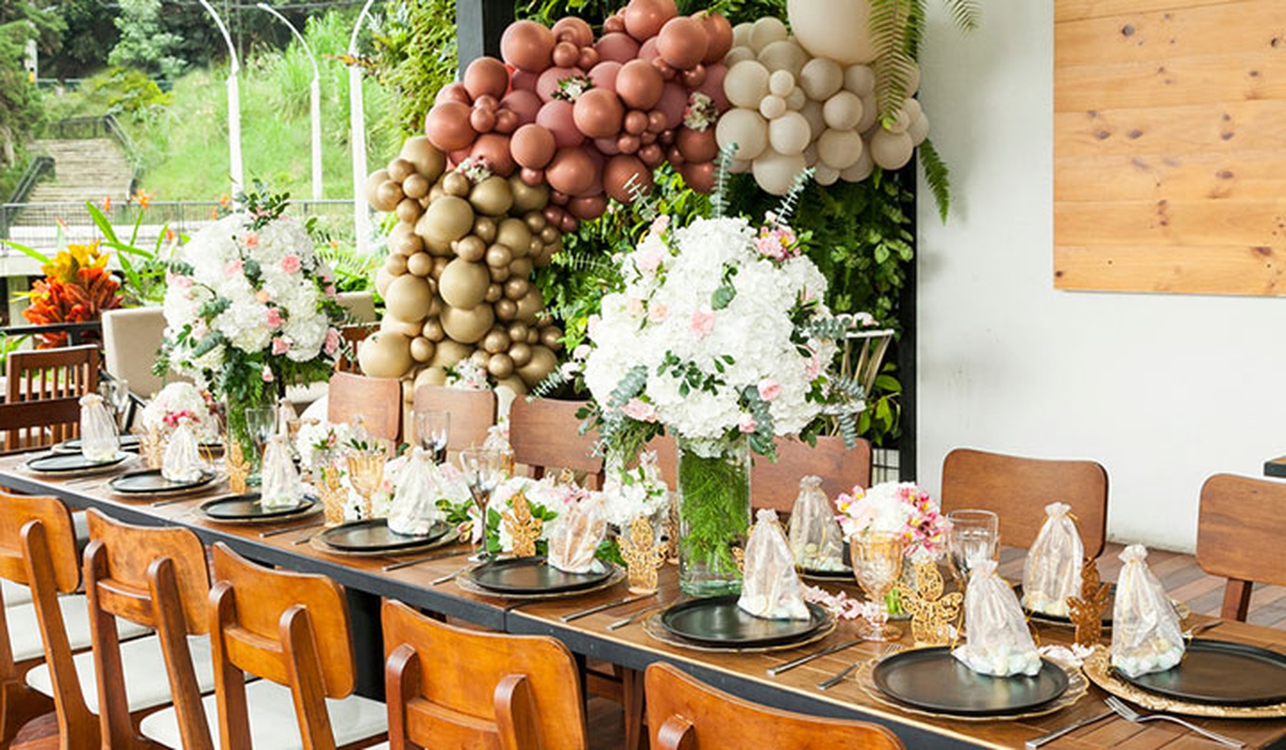 Mesa decorada de comunión. Hay platos negros, ramos de flores rosas y un arco de globos rosas, beige y blancos