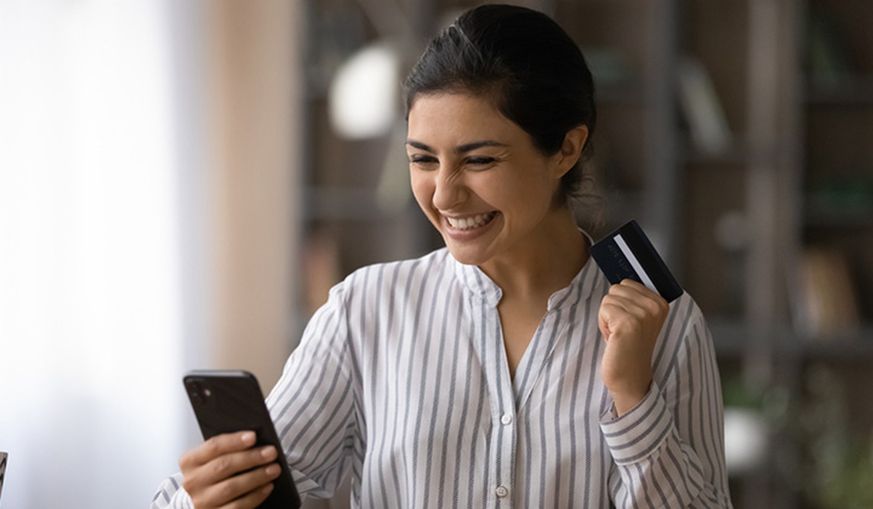 Mujer joven muy sonriente mirando su smartphone y con una tarjeta de crédito en la otra mano