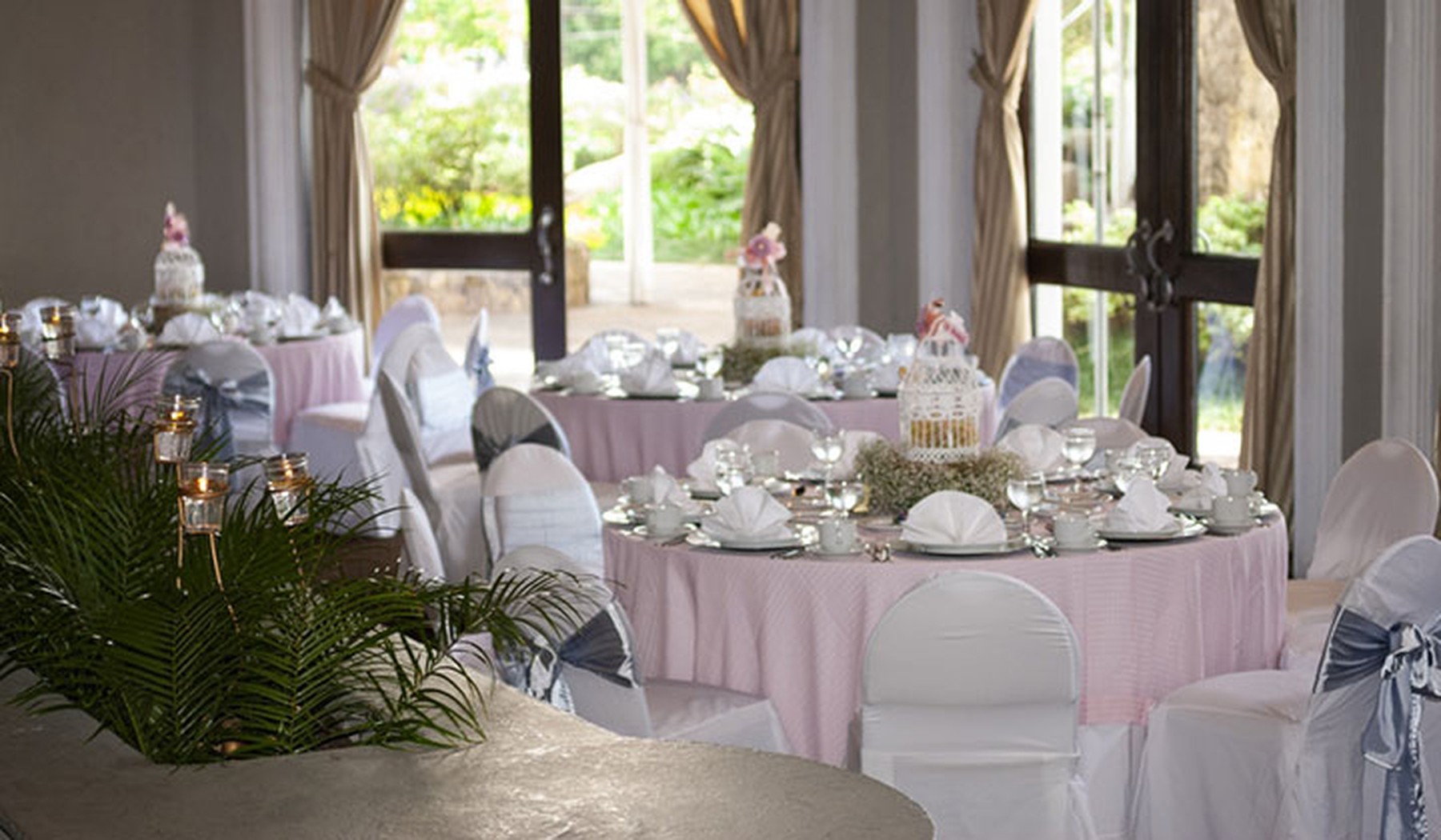 Tres mesas redondas decoradas de comunión con manteles rosas claritos. En primer plano hay una planta de hojas verdes y al fondo dos puertas de cristal.
