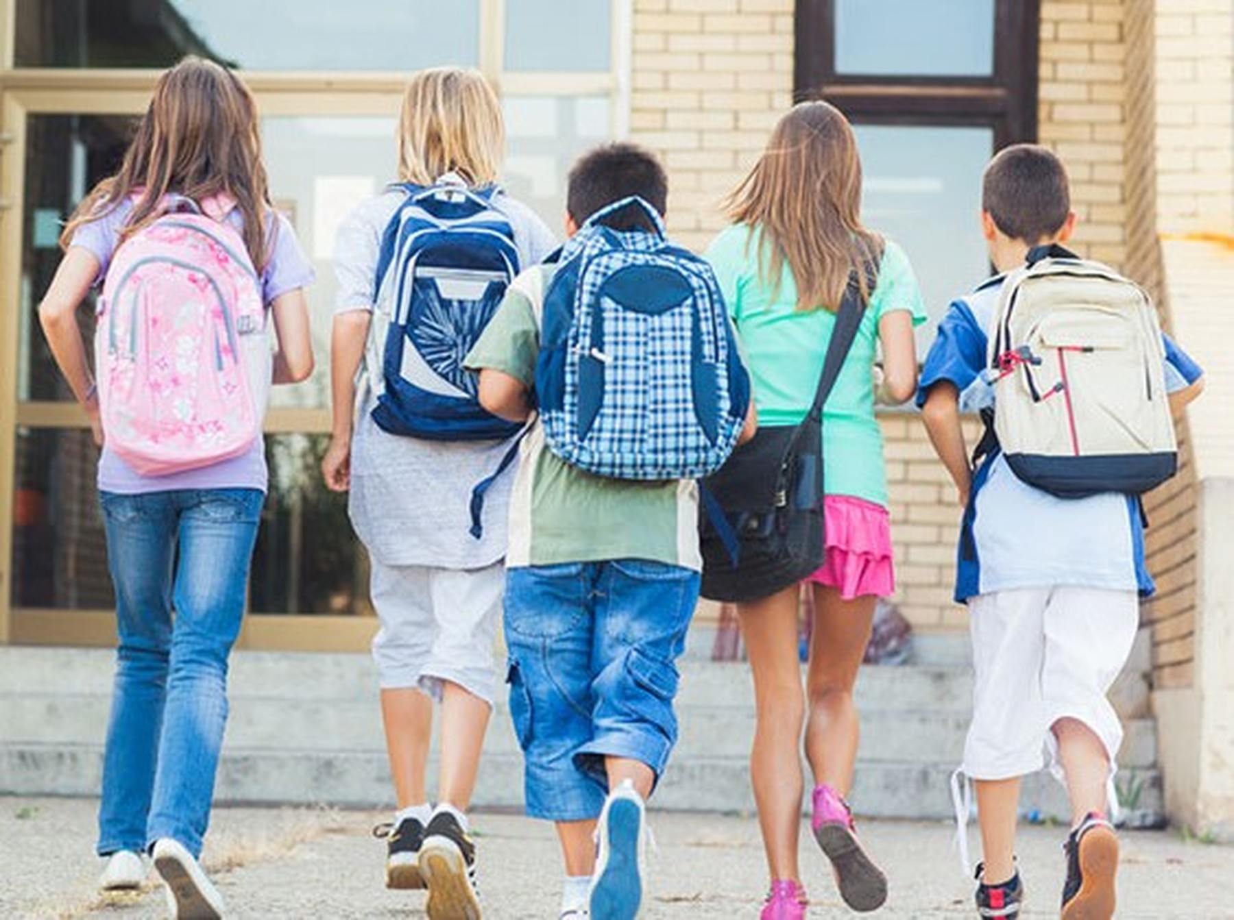 5 niños y niñas de espaldas caminando hacia la puerta de su colegio con mochilas en las espaldas