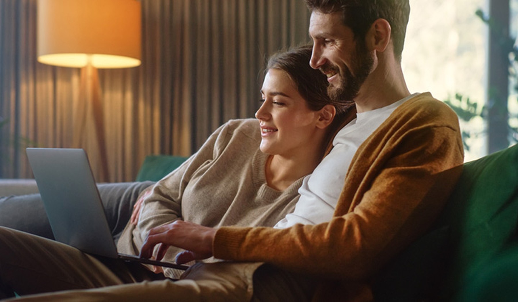 Pareja de hombre y mujer tumbados en un sofá verde abrazados mientras miran la pantalla de un portátil