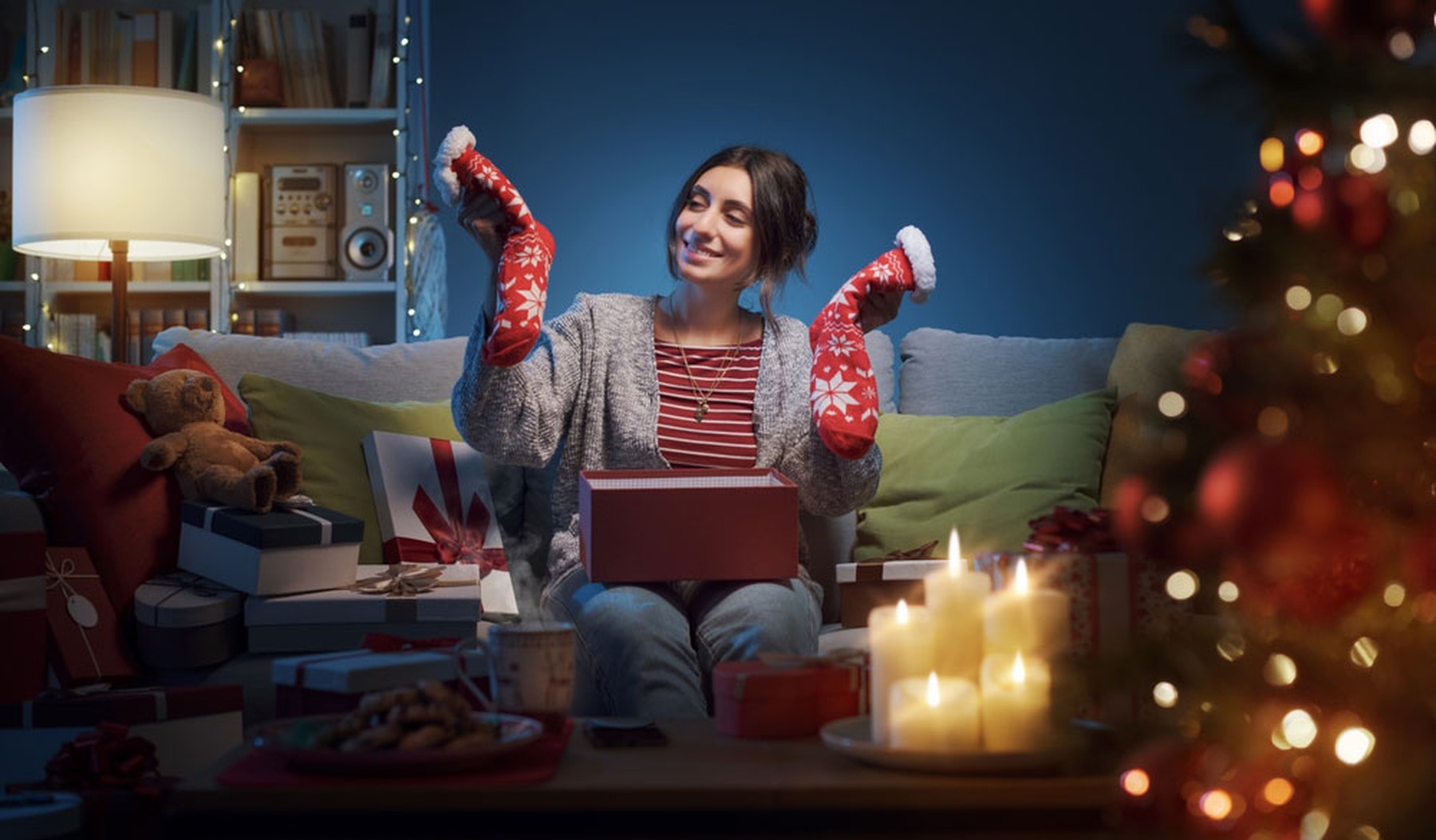 Mujer feliz sentada en el sofá de casa y abriendo regalos de Navidad, tiene en las manos unos calcetines rojos navideños.