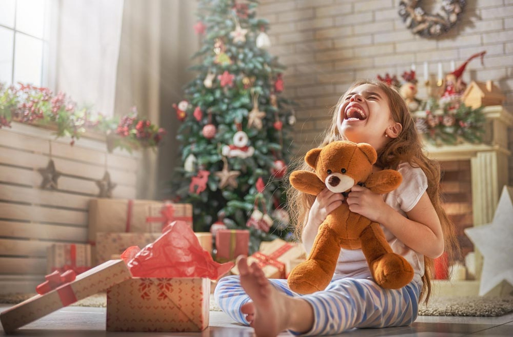 Una niña pequeña abriendo regalo cerca del árbol de Navidad con un peluche de un osito en las manos.