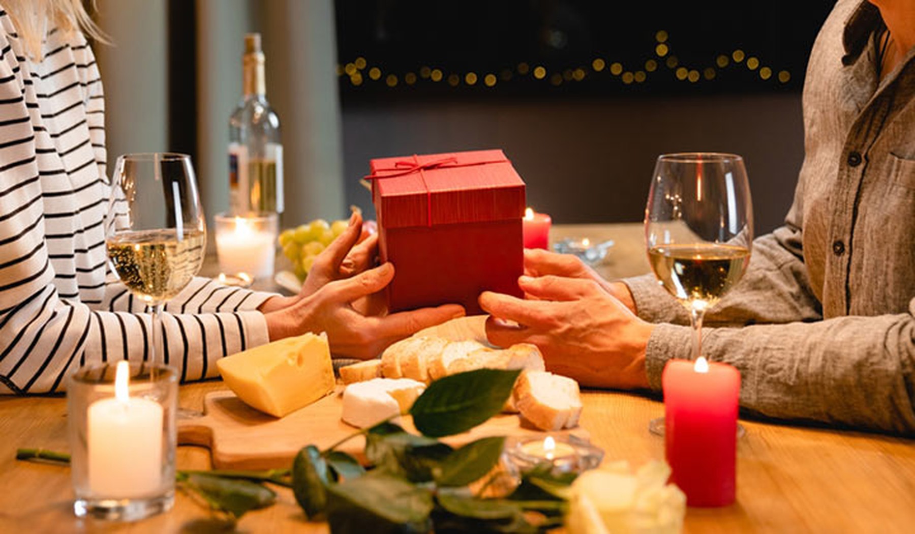Foto de las manos de una pareja dándose regalos por su aniversario mientras disfrutan de una cena romántica con quesos y vino.