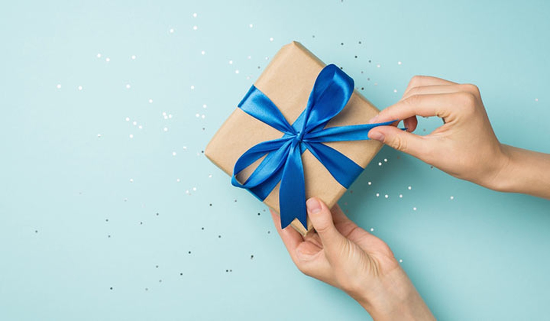 Foto en vista superior en primera persona de manos desenvolviendo una caja de regalo con un lazo de satén azul sobre lentejuelas brillantes en un fondo de color azul pastel con espacio en blanco.