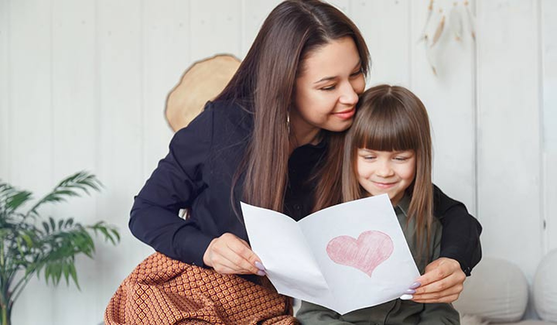 Una hija sentada con su madre dándole una tarjeta con un corazón por el Día de la Madre
