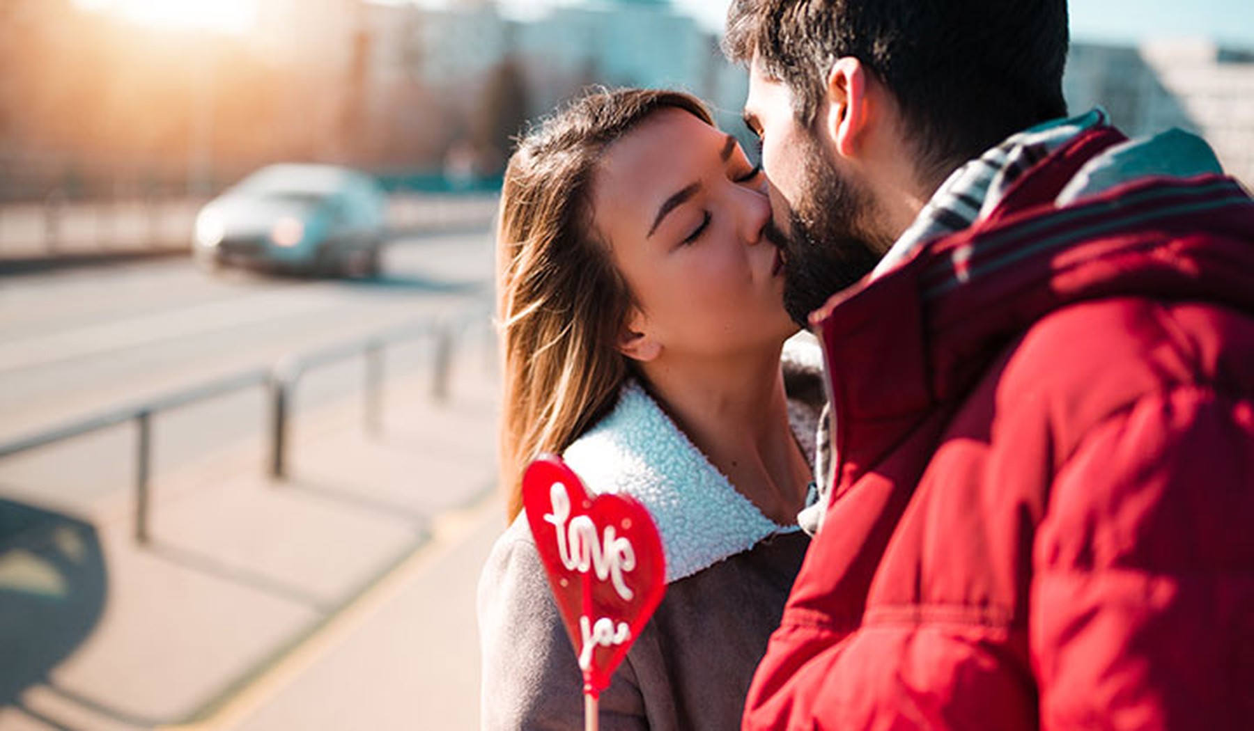 Pareja joven dándose un beso mientras él sostiene una piruleta roja en forma de corazón que pone Love you