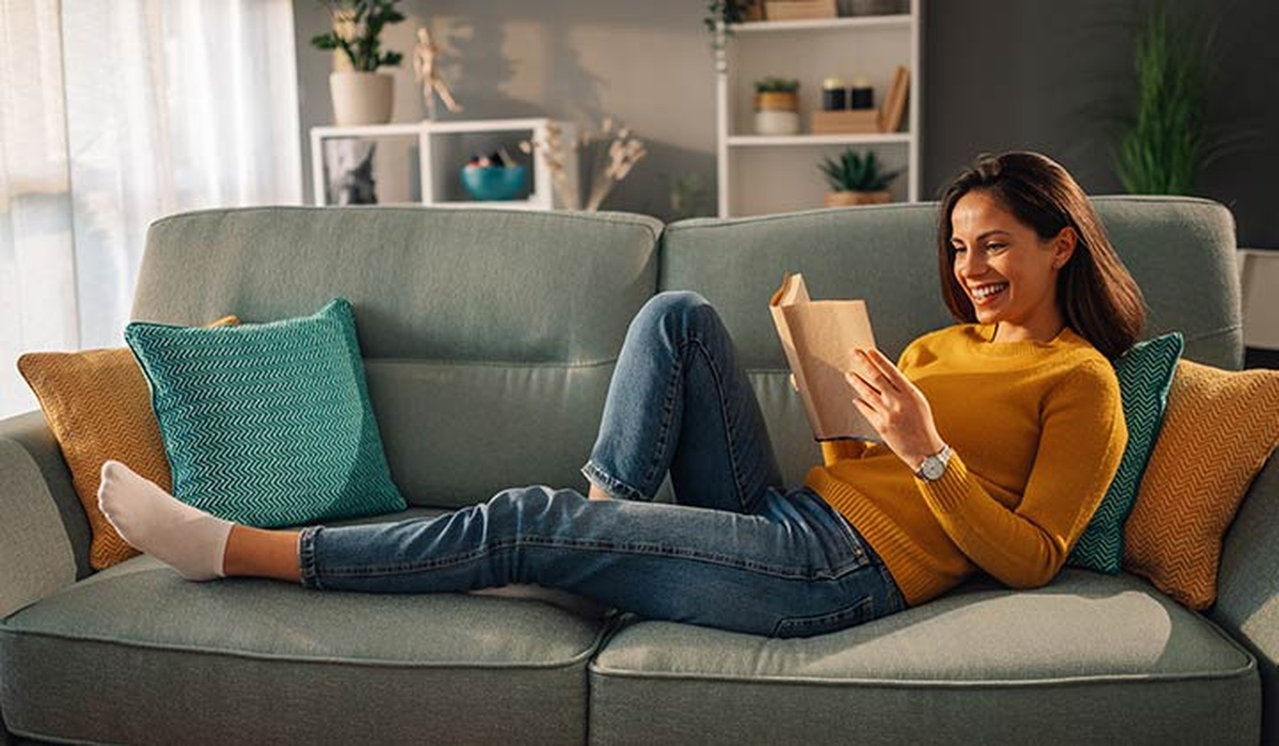 Mujer sonriente tumbada en el sofá de su casa mientras lee un libro