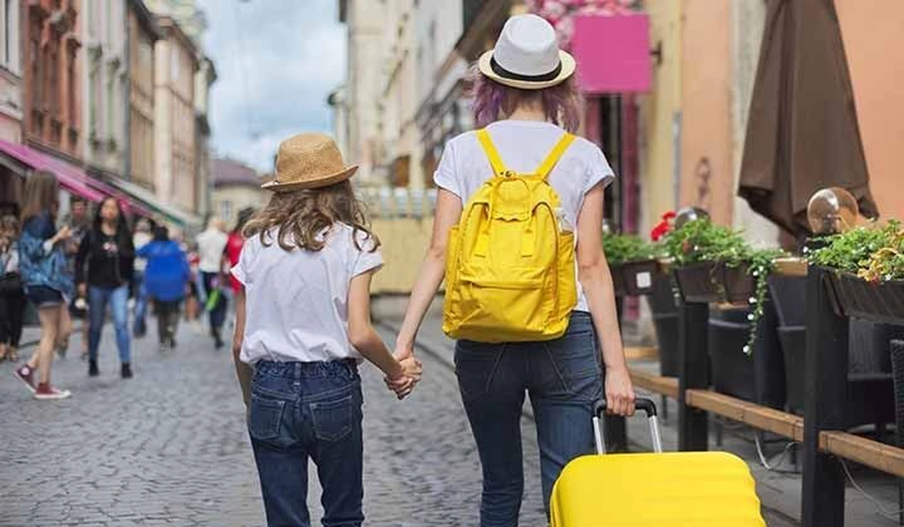 Vista posterior de una mujer joven con su hija de la mano llevando una maleta de ruedas amarilla y una mochila a juego mientras pasean haciendo turismo por una ciudad