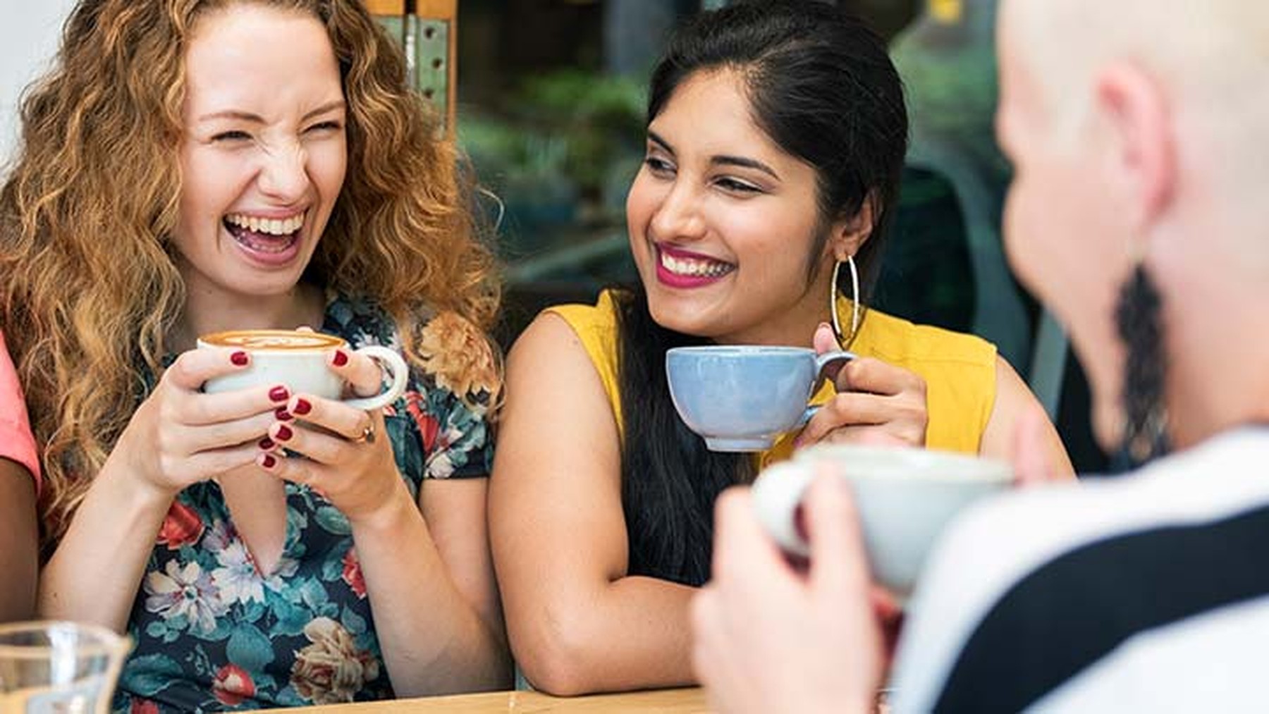 Tres mujeres jóvenes bebiendo café en diferentes tazas mientras hablan y se ríen.