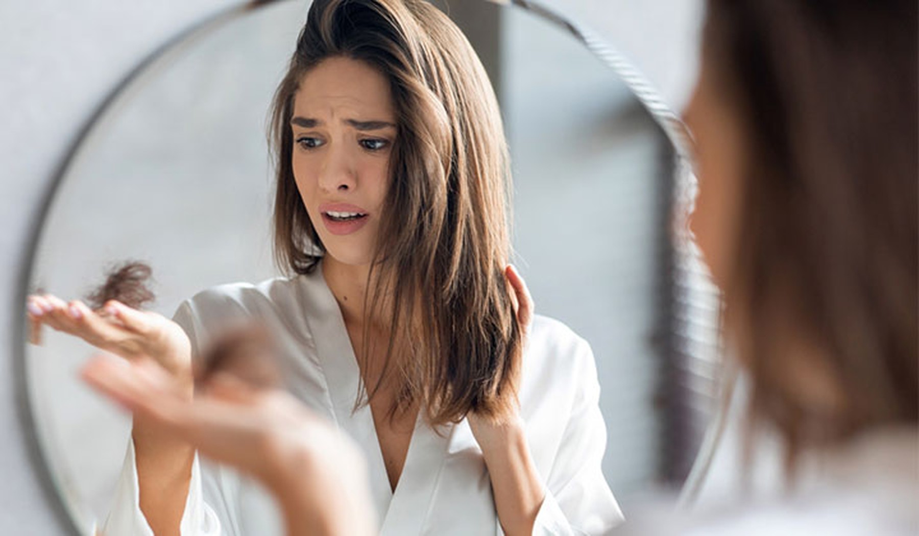 Mujer triste mirando un mechón de pelo en su mano frente a un espejo