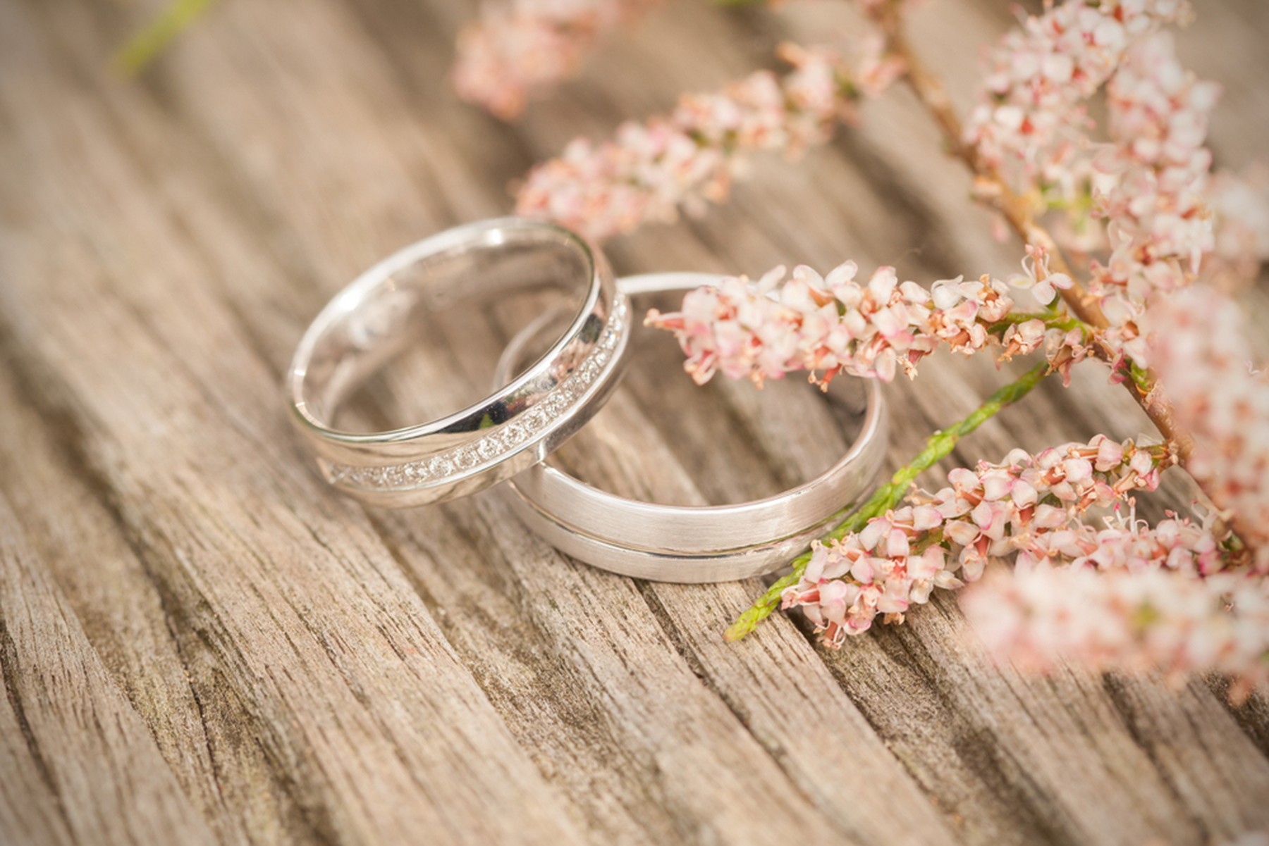 Dos alianzas de plata u oro blanco con un ramito de flores rosas al lado. Todo está sobre una mesa de madera.