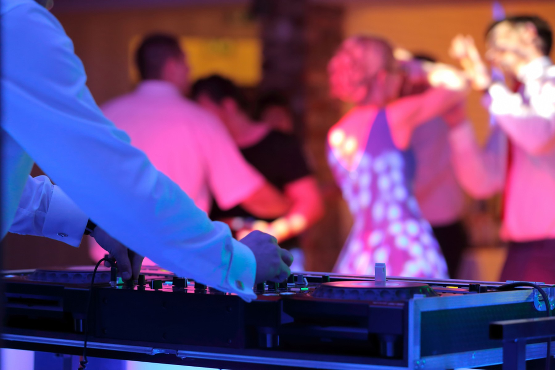 DJ con su mesa de mezclas iluminado por una luz azul. En el fondo se ve a gente bailar iluminados por luces rosas.