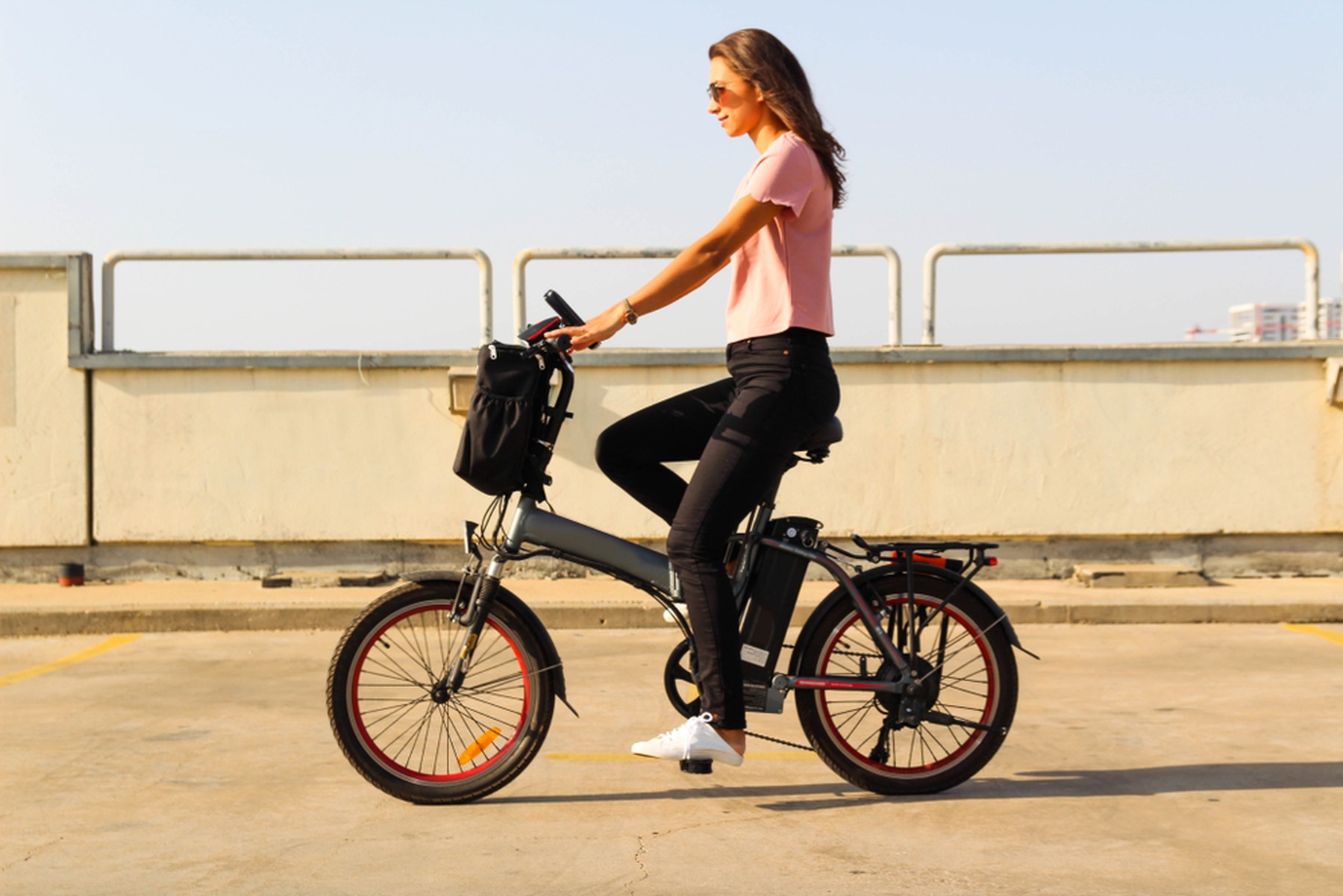 Mujer joven con una camiseta de manga corta rosa y unos vaqueros negros montando en una bicicleta eléctrica por la ciudad