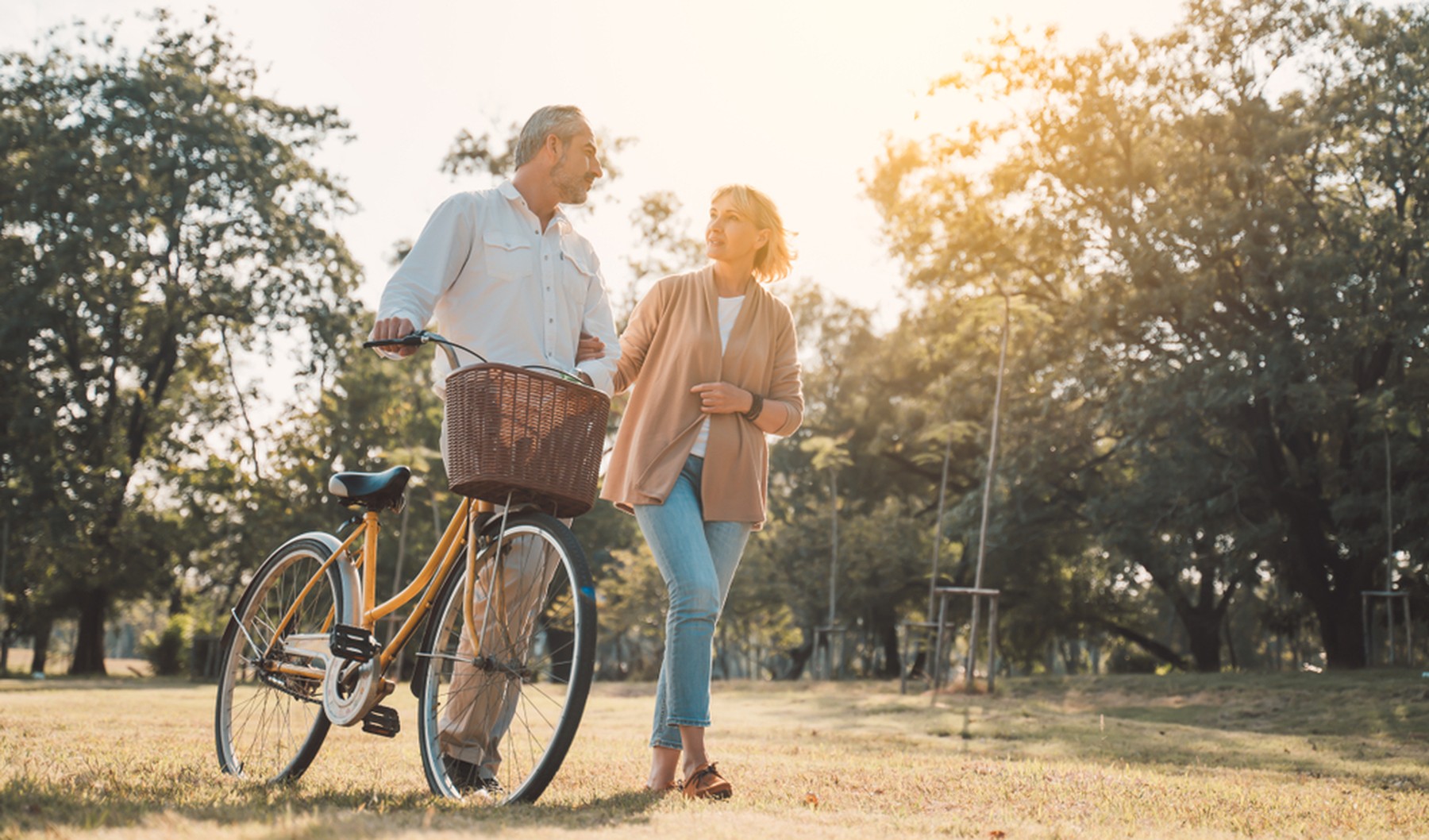 Hombre con una bicicleta de paseo amarilla con cesta caminando por un parque. Del brazo va cogida una mujer con el pelo corto rubio