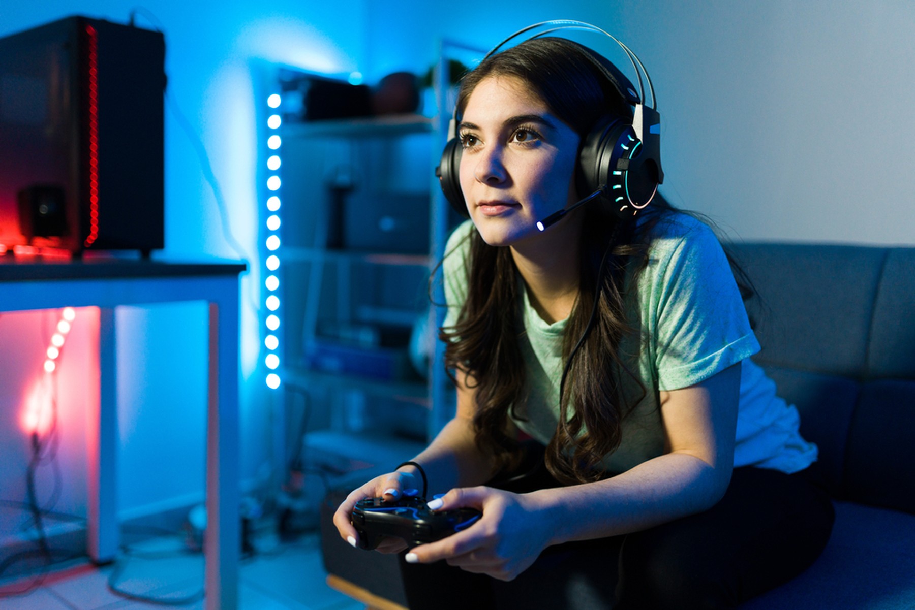 Chica joven con el pelo largo sentada en un sofá jugando a videojuegos. Lleva unos auriculares de gamer con micrófono y de fondo se ven luces led azules y rojas