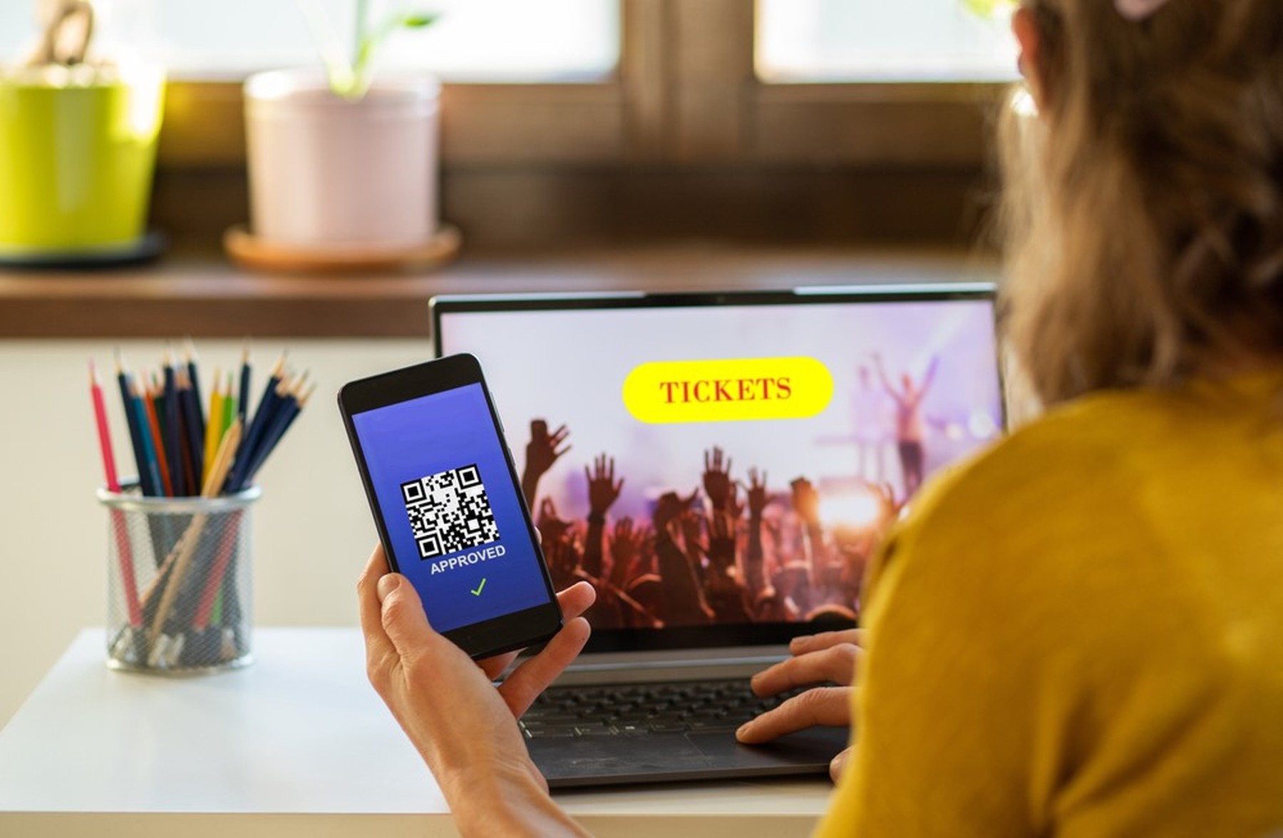 Mujer mirando tickets para un concierto en su portátil. Con una mano sostiene un smartphone donde se ve un código QR con el fondo azul.