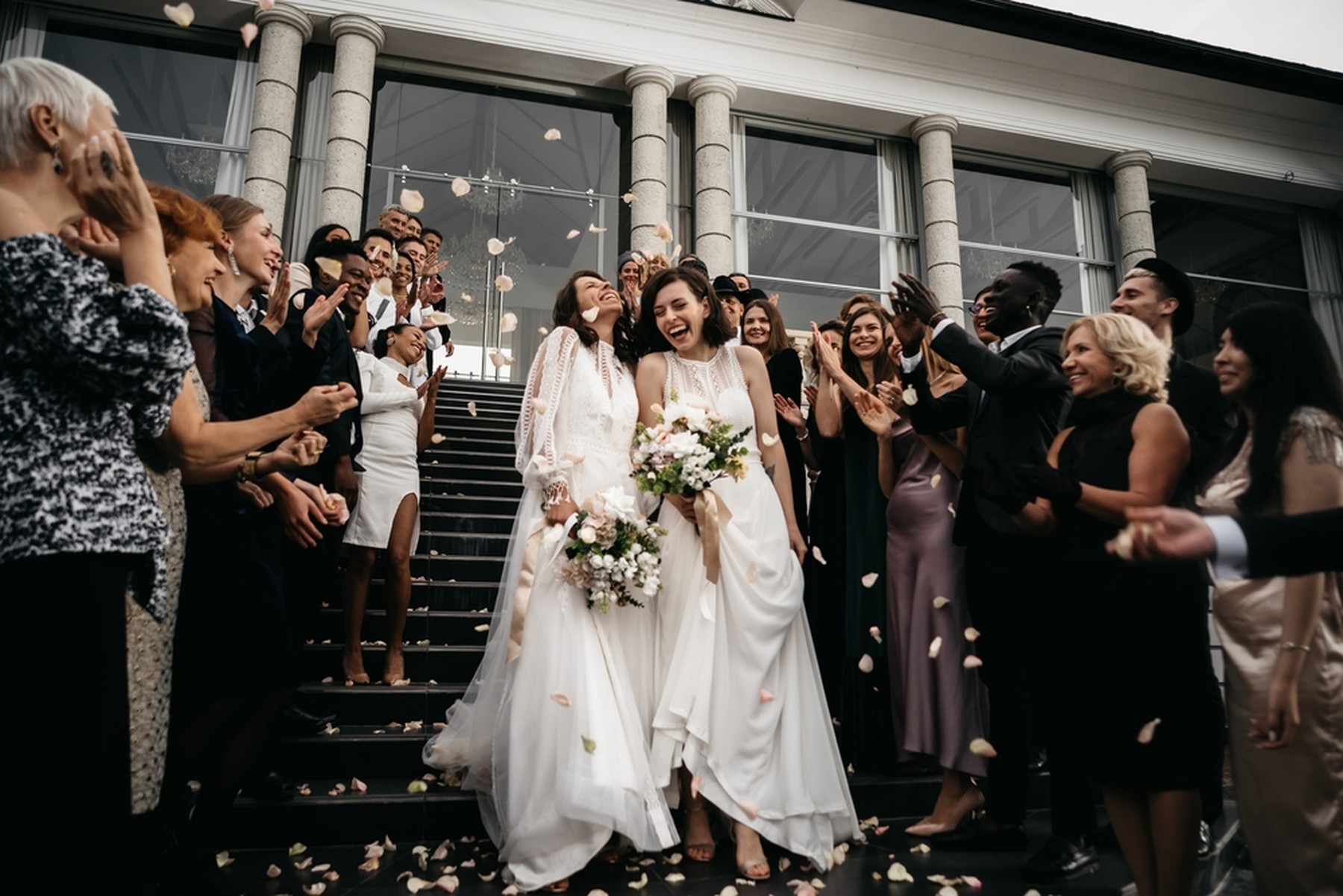 Dos mujeres jóvenes vestidas de novia saliendo de un edificio tras casarse. Los invitados les están lanzando pétalos de rosas rosas.