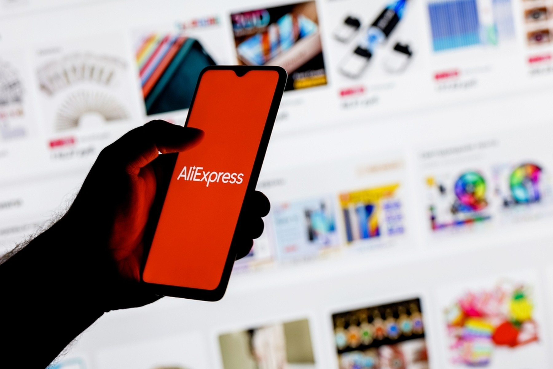 Mano de hombre con un smartphone con la pantalla en rojo con el logo de AliExpress, de fondo se ve la web de AliExpress