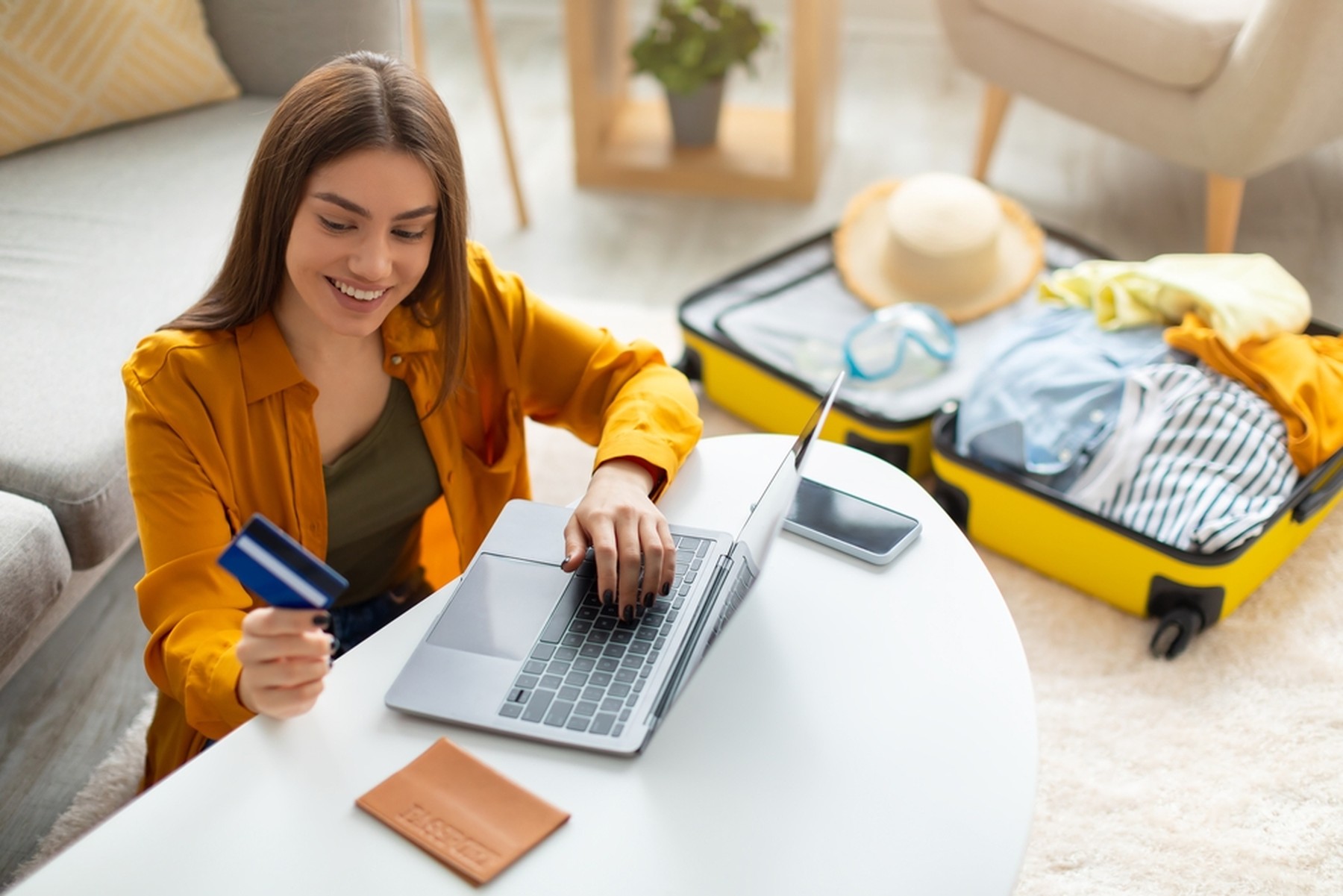 Mujer joven sonriendo mientras mira su tarjeta de crédito y utiliza un ordenador portátil. En la mesa tiene una libreta y al fondo se ve una maleta amarilla abierta.