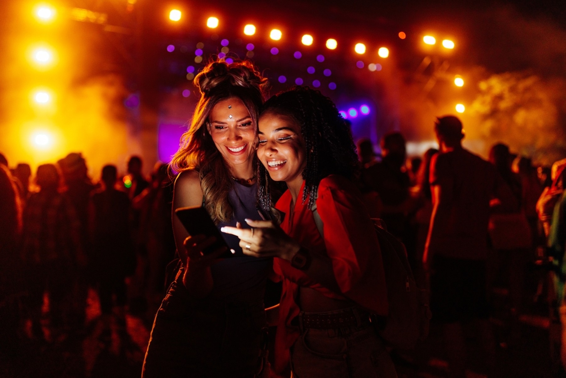 Dos chicas jóvenes mirando un smartphone en un festival. De fondo se ven las luces del escenario. Ellas llevan maquillaje festivo.