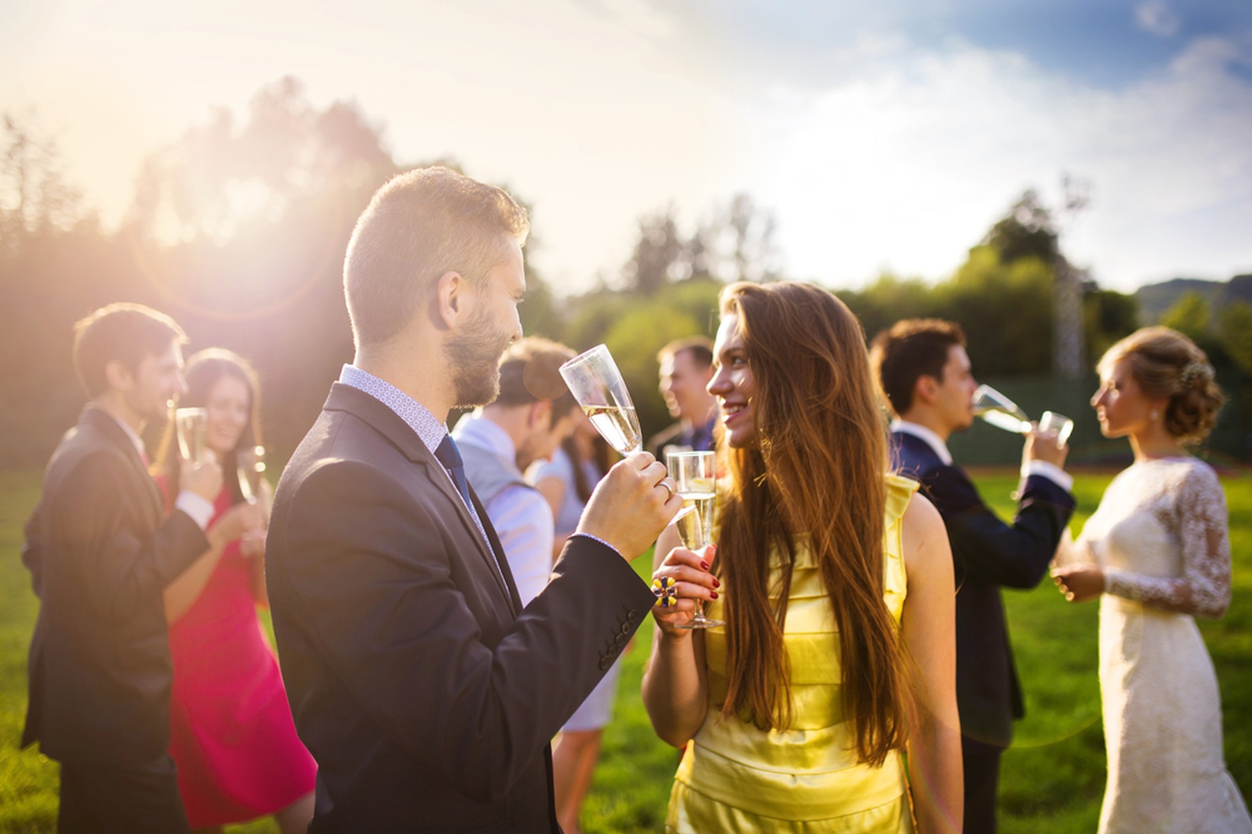 Una pareja en primer plano brindando con champán en una boda. En el fondo se ve a los novios y otros invitados