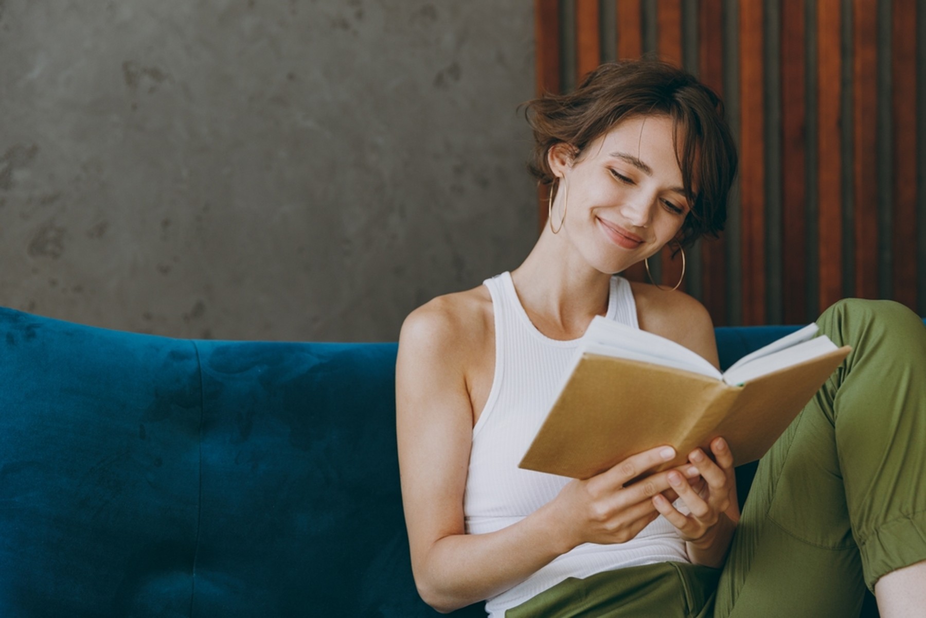 Mujer joven con el pelo corto leyendo un libro con la portada marrón. Está sentada en un sofá de terciopelo azul y sonríe.