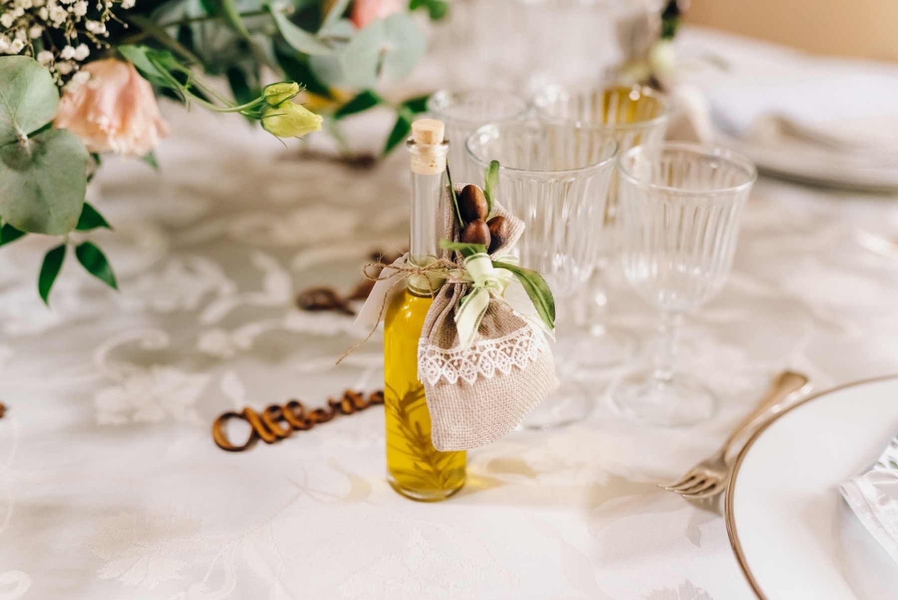 Botellita de aceite con un adorno de tela y plantas. Está encima de una mesa con mantel blanco, copas de cristal y flores