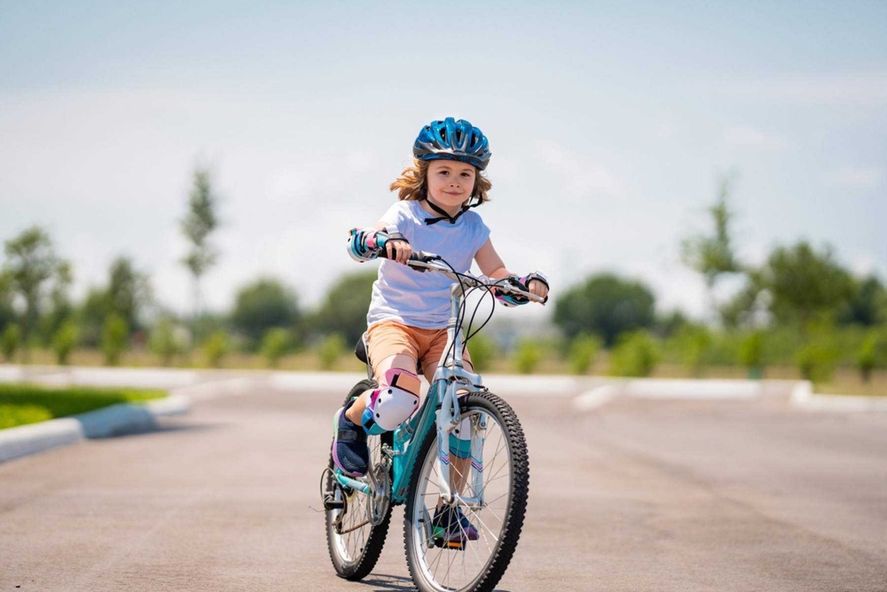 Niño montando en una bicicleta azul en una zona de asfalto en un parque. El niño lleva un casco azul, coderas y rodilleras